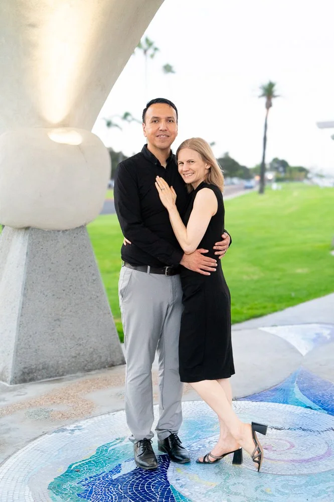 A couple standing close together outdoors, smiling. The woman is wearing a black dress and heels, the man is dressed in a black shirt and light gray pants. They are standing on a circular mosaic design with a colorful pattern, near a modern concrete