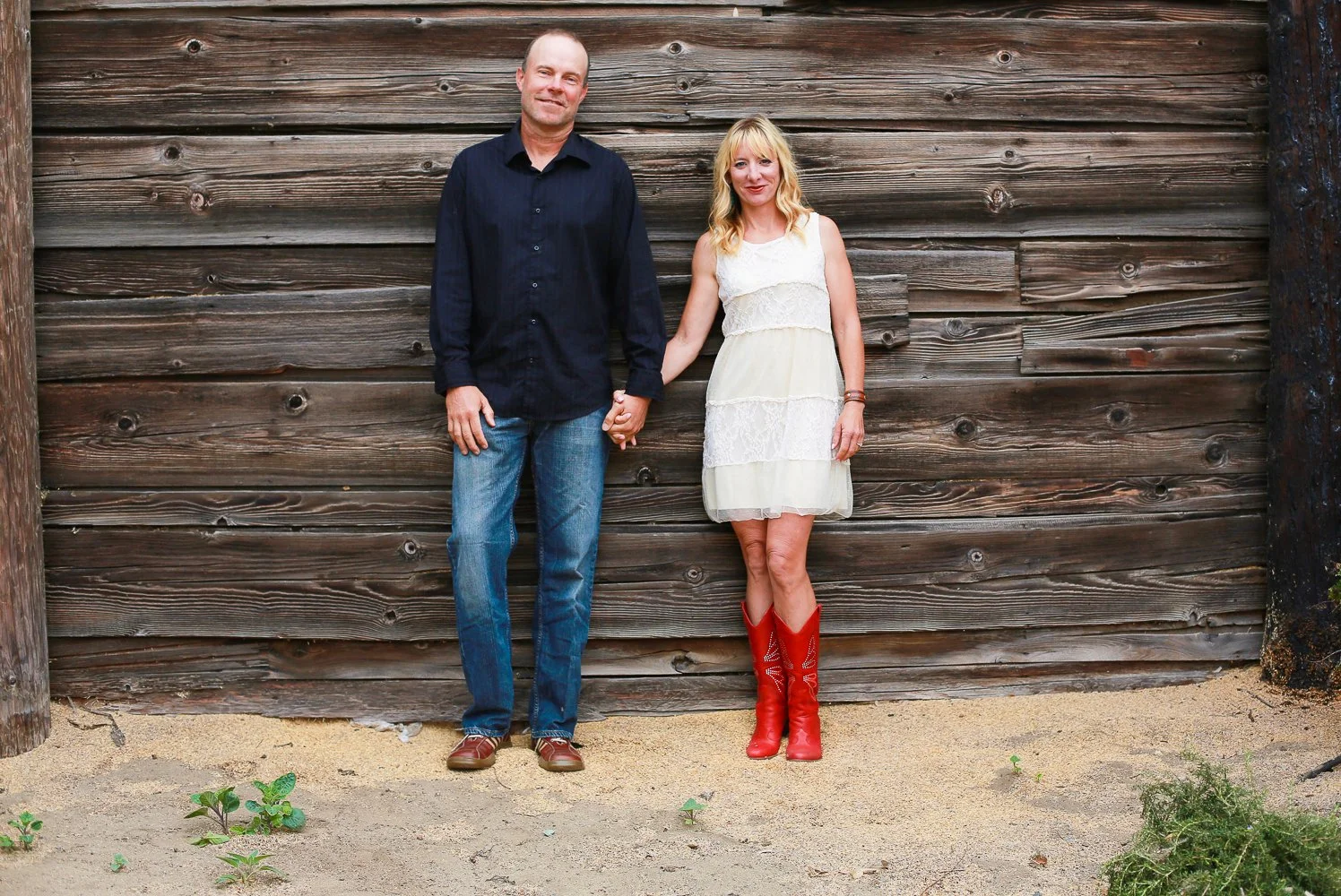 A man and woman holding hands, standing in front of a wooden wall, with some plants and sand on the ground.