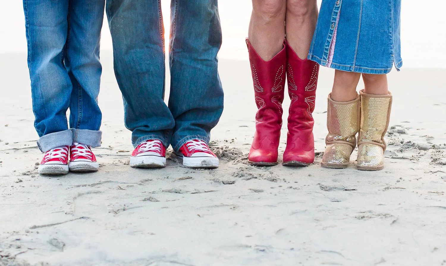 Close-up of four people's legs and feet standing on sandy ground, wearing colorful shoes and boots.