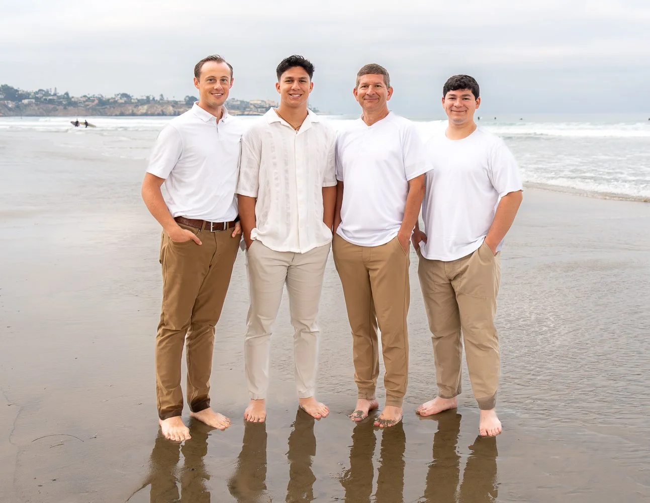 Four men standing on the beach near the water, smiling at the camera. They are wearing white shirts and khaki pants, and are barefoot, with the ocean and cloudy sky in the background.