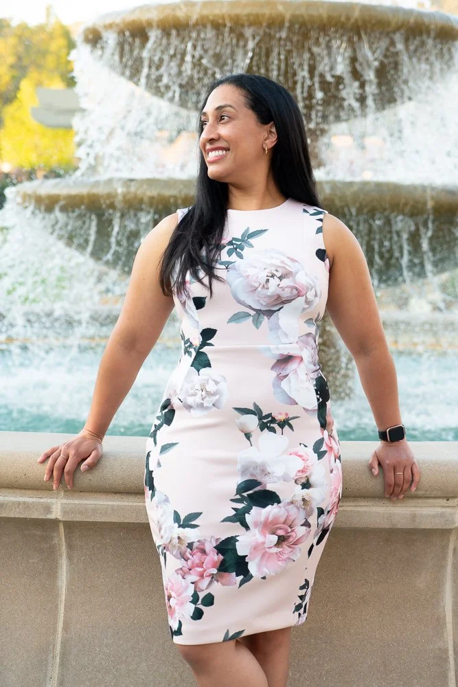 A woman in a white floral dress standing by a fountain with water cascading in the background, smiling and looking to her left.