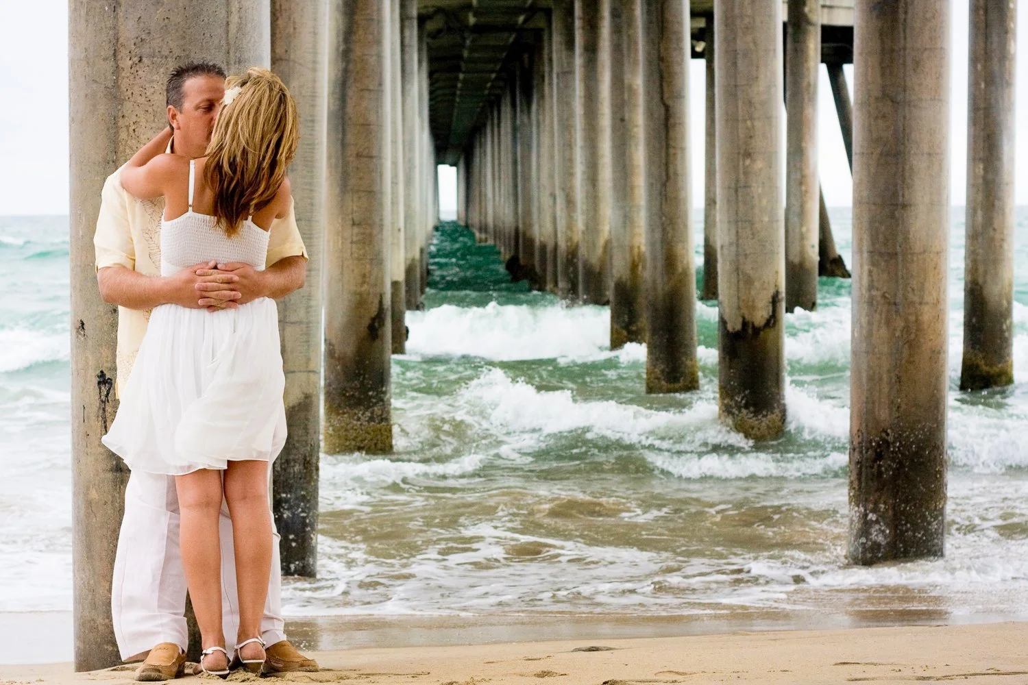 A couple embracing on a beach near a pier, with ocean waves crashing against the pilings.