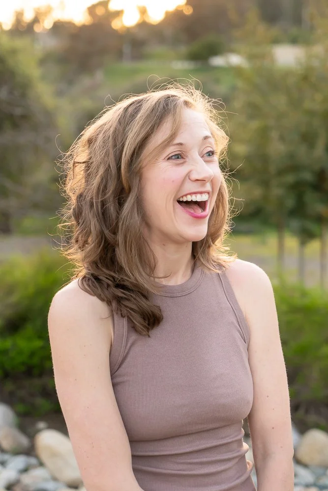 A young woman with light brown, shoulder-length wavy hair, smiling and laughing outdoors during sunset, wearing a sleeveless beige top, with trees and rocks in the background.
