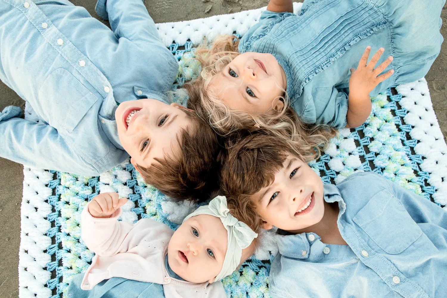 Four children lying on a blanket, smiling and looking up, outdoors with a concrete surface nearby.