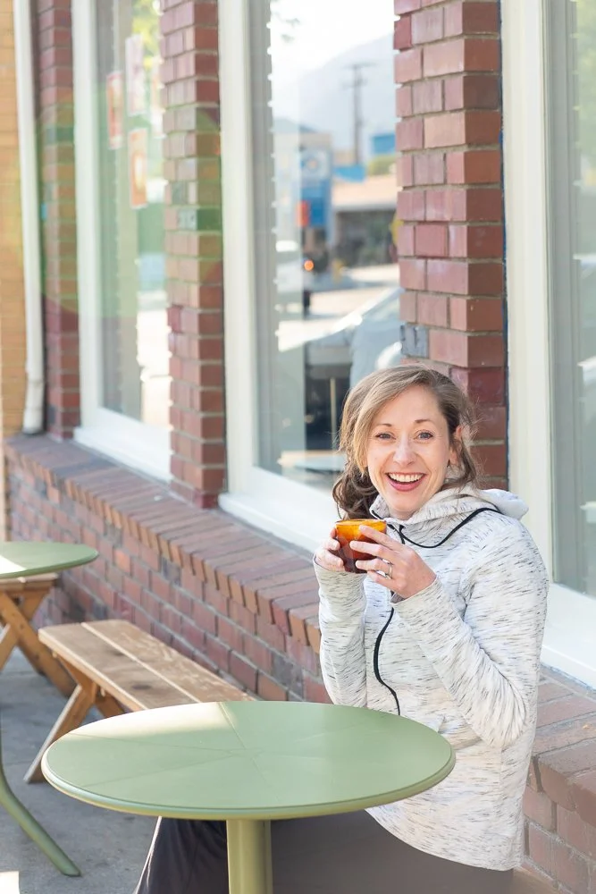 A woman sitting outside at a green table, holding a drink and smiling, in front of a brick building with large windows.