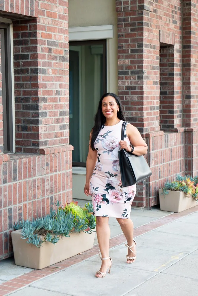 Woman in floral dress walking on sidewalk carrying a black bag in front of a brick building with flower pots.