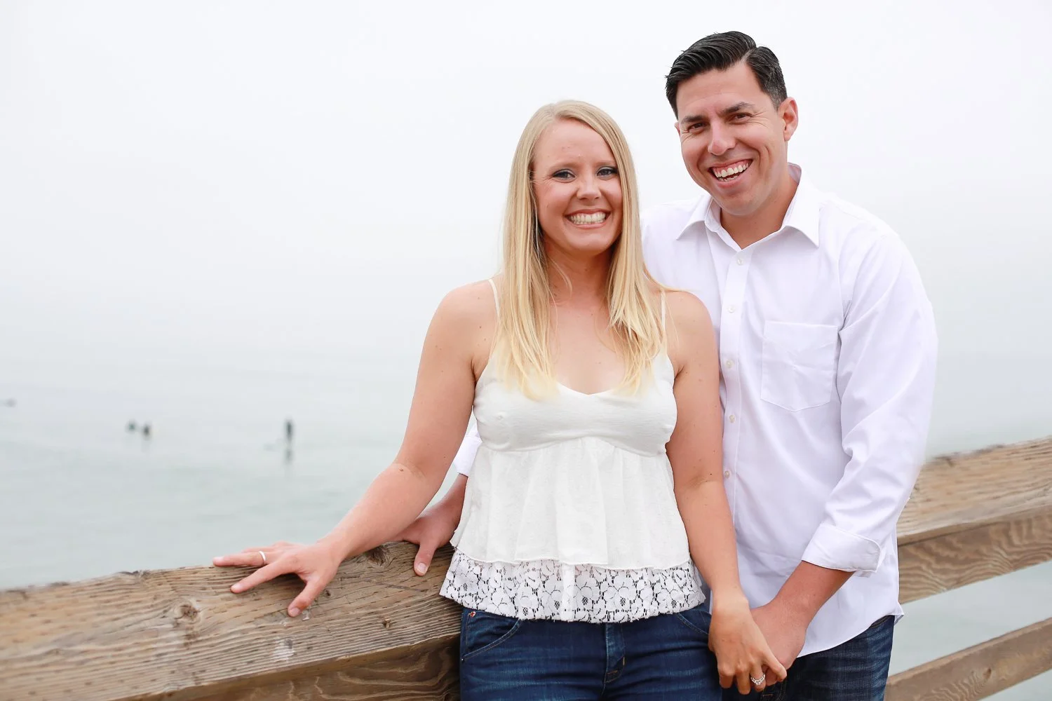 A smiling couple on a wooden pier, with the woman in a white sleeveless top and the man in a white button-up shirt, standing by the ocean in a foggy setting.