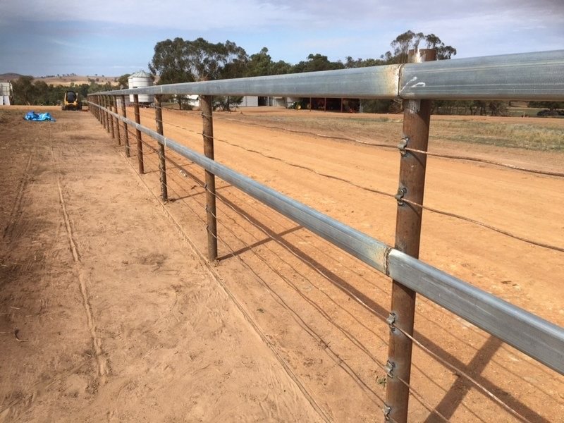 A wide view of a rural farm field with a metal and wooden fence in the foreground, dirt path alongside, and farm equipment and trees in the background.