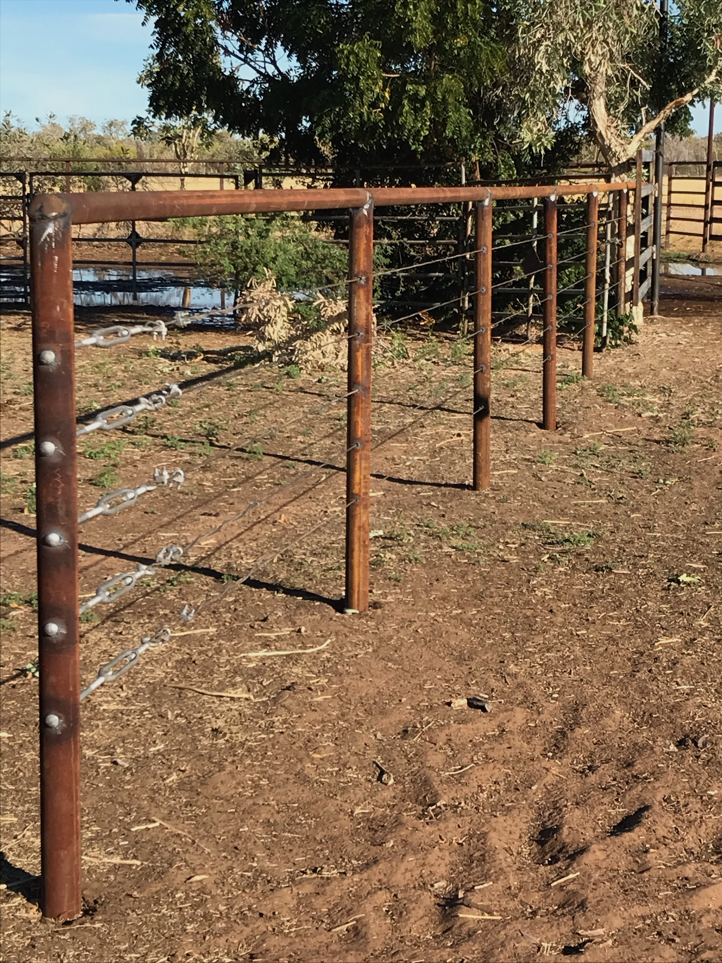 Empty outdoor playground with rusty metal climbing structure on dirt ground, surrounded by trees and a fence.
