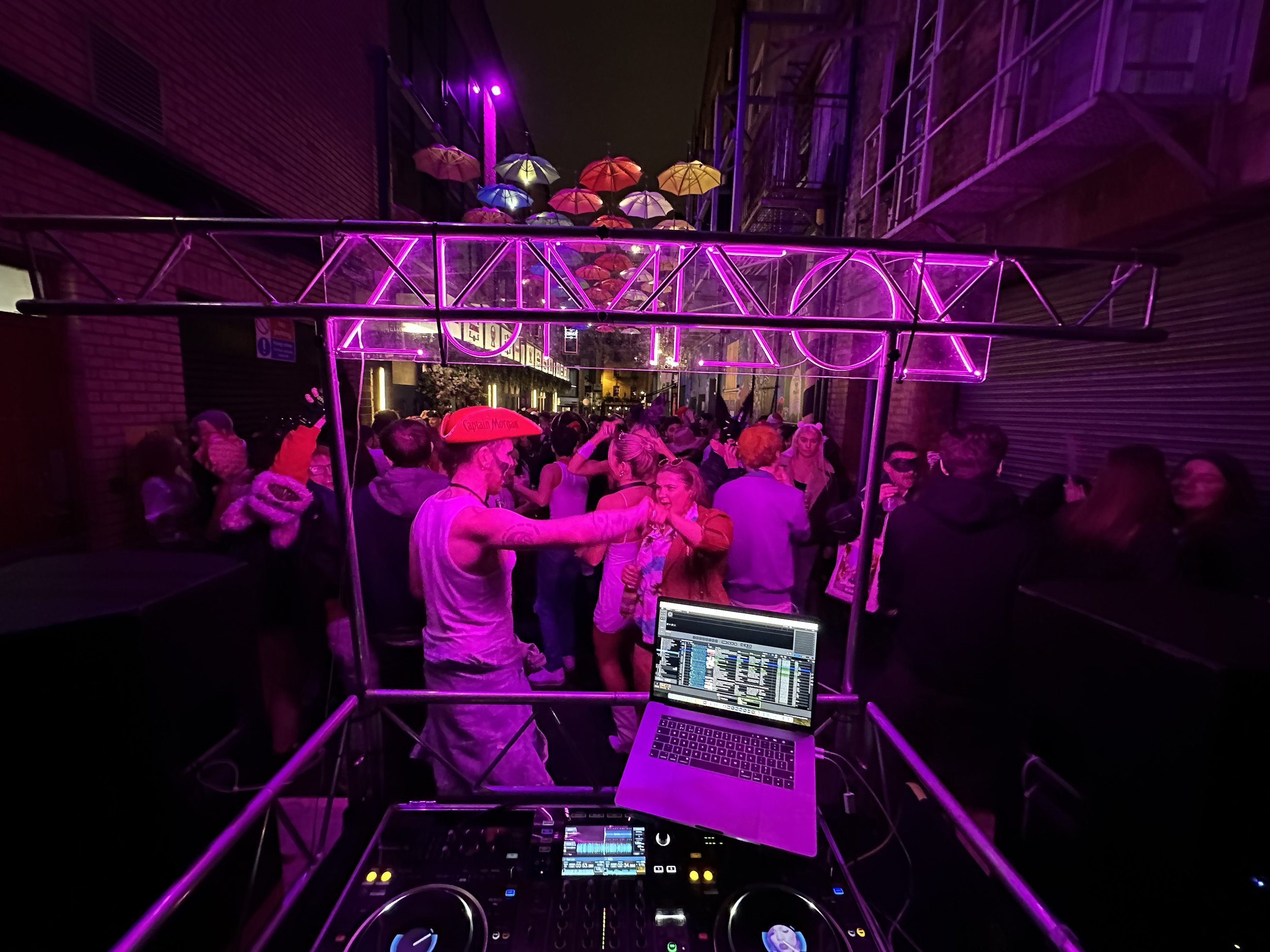 Nightclub scene with a DJ setup in the foreground and a crowded dance floor with people dancing and socializing under purple and pink lighting.