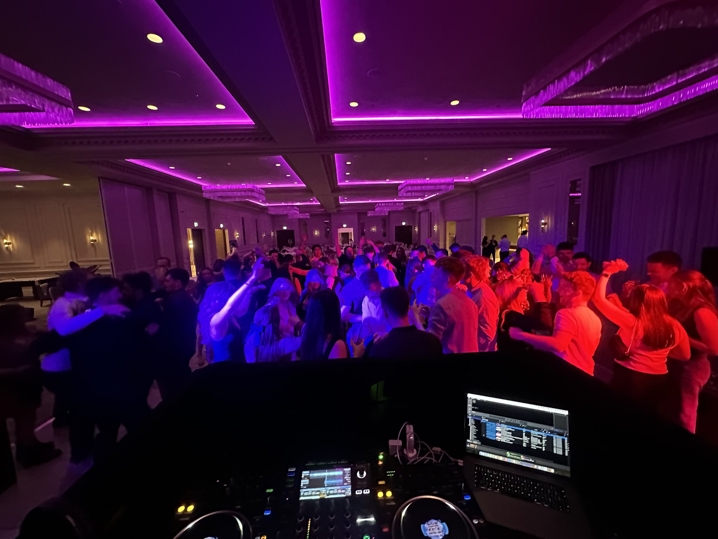Crowd of people dancing at a party in a large, decorated ballroom with purple and pink lighting.