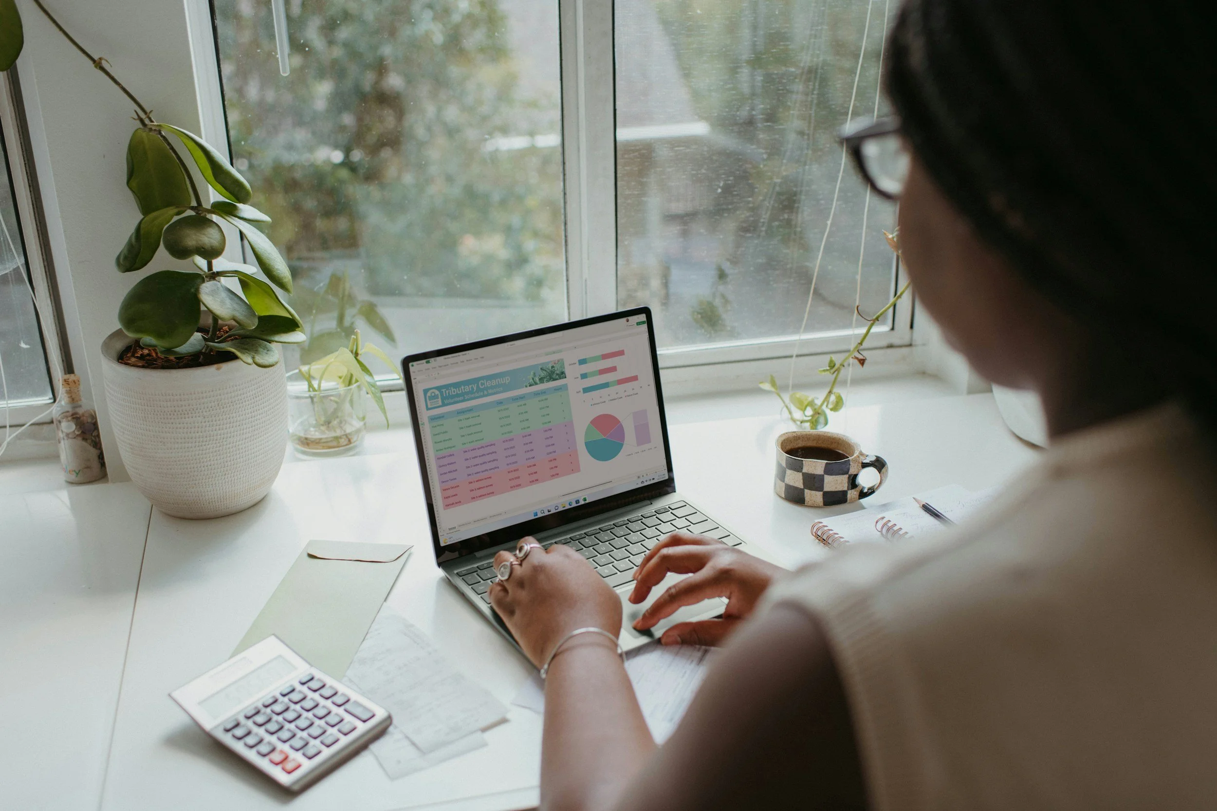 Person working on a laptop at a white desk with a calculator, papers, and a mug, near a window with houseplants.
