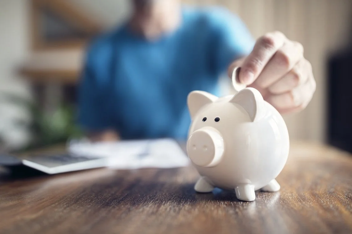 Person placing a coin into a white piggy bank on a wooden table.