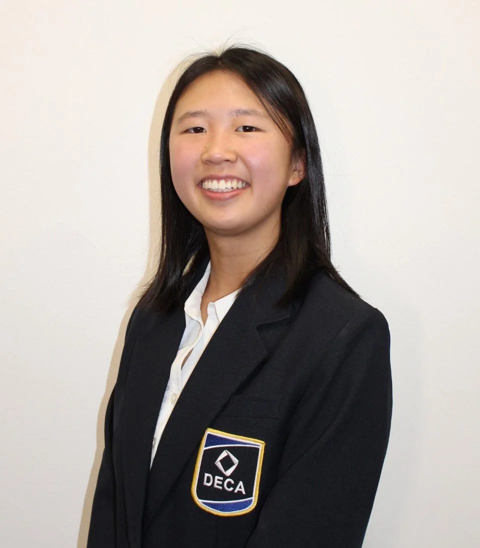 Young woman with black hair wearing a black blazer with a DECA patch on the chest, standing against a plain white wall.