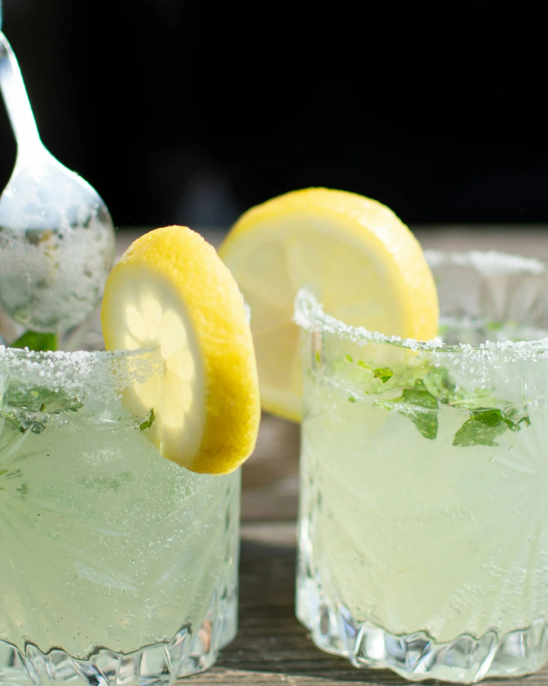 Two glasses of lemon cocktails with salted rims, lemon slices, and mint leaves, with lemon slices in the background on a wooden surface.