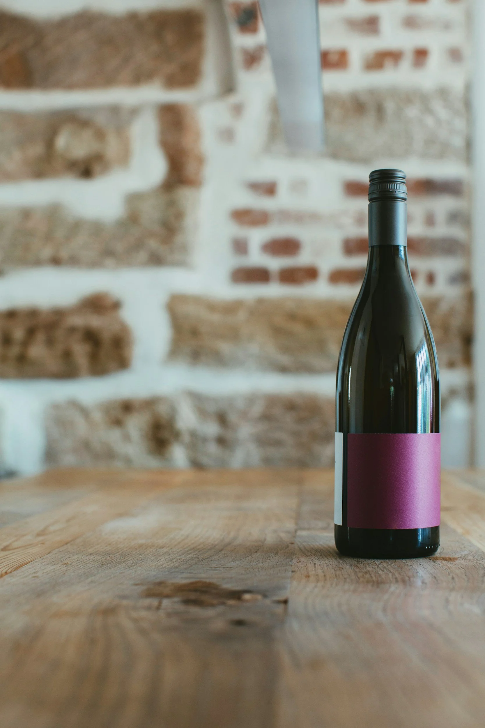 A dark glass bottle with a pink label sitting on a wooden table with a brick wall in the background.