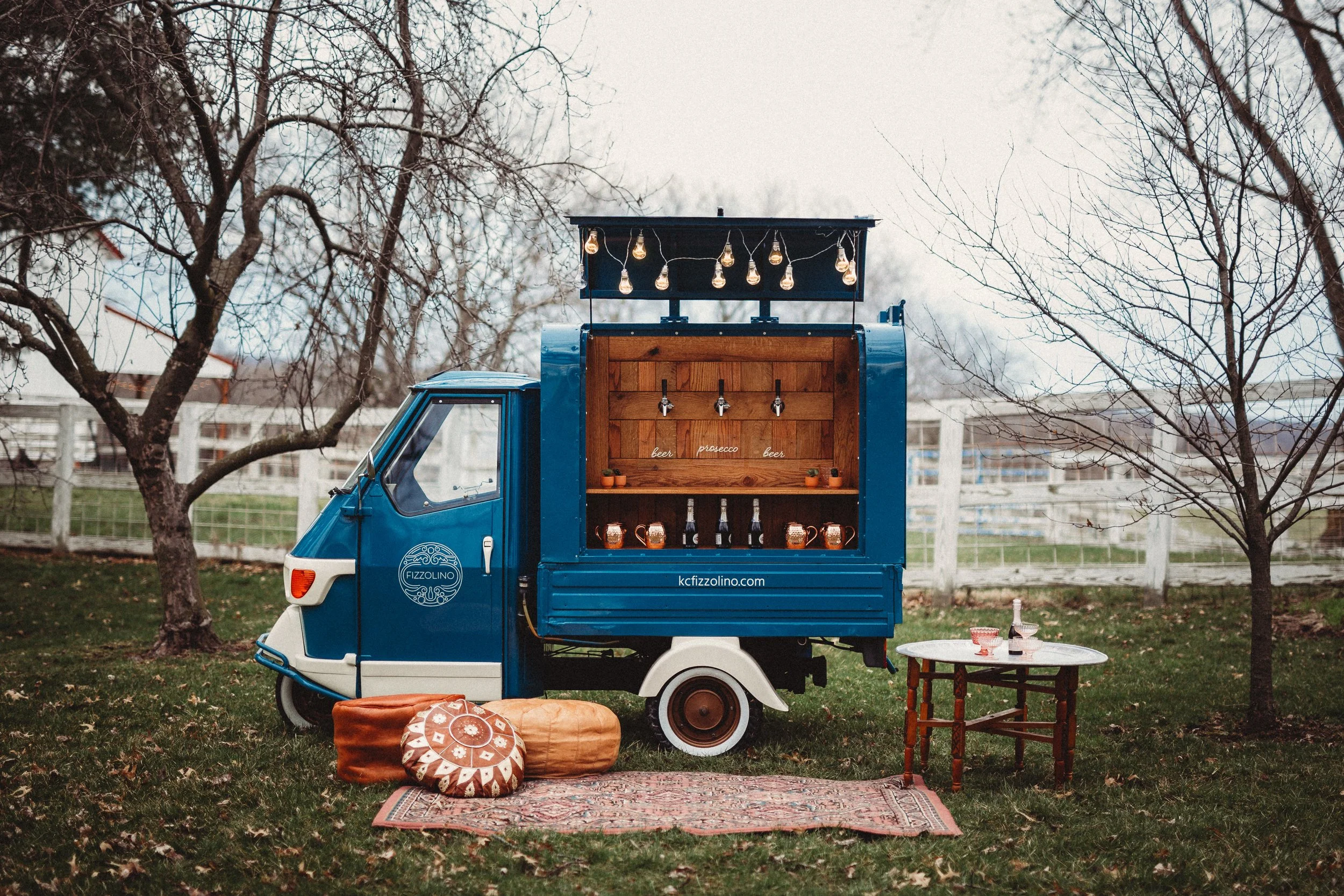 A vintage blue Piaggio Ape mini truck converted into a mobile bar with a wooden bar interior, string lights hanging above, and bottles and glasses on the shelves. Outdoor setting with leafless trees, a rug on the grass, and a small round table with wine glasses and a bottle nearby.