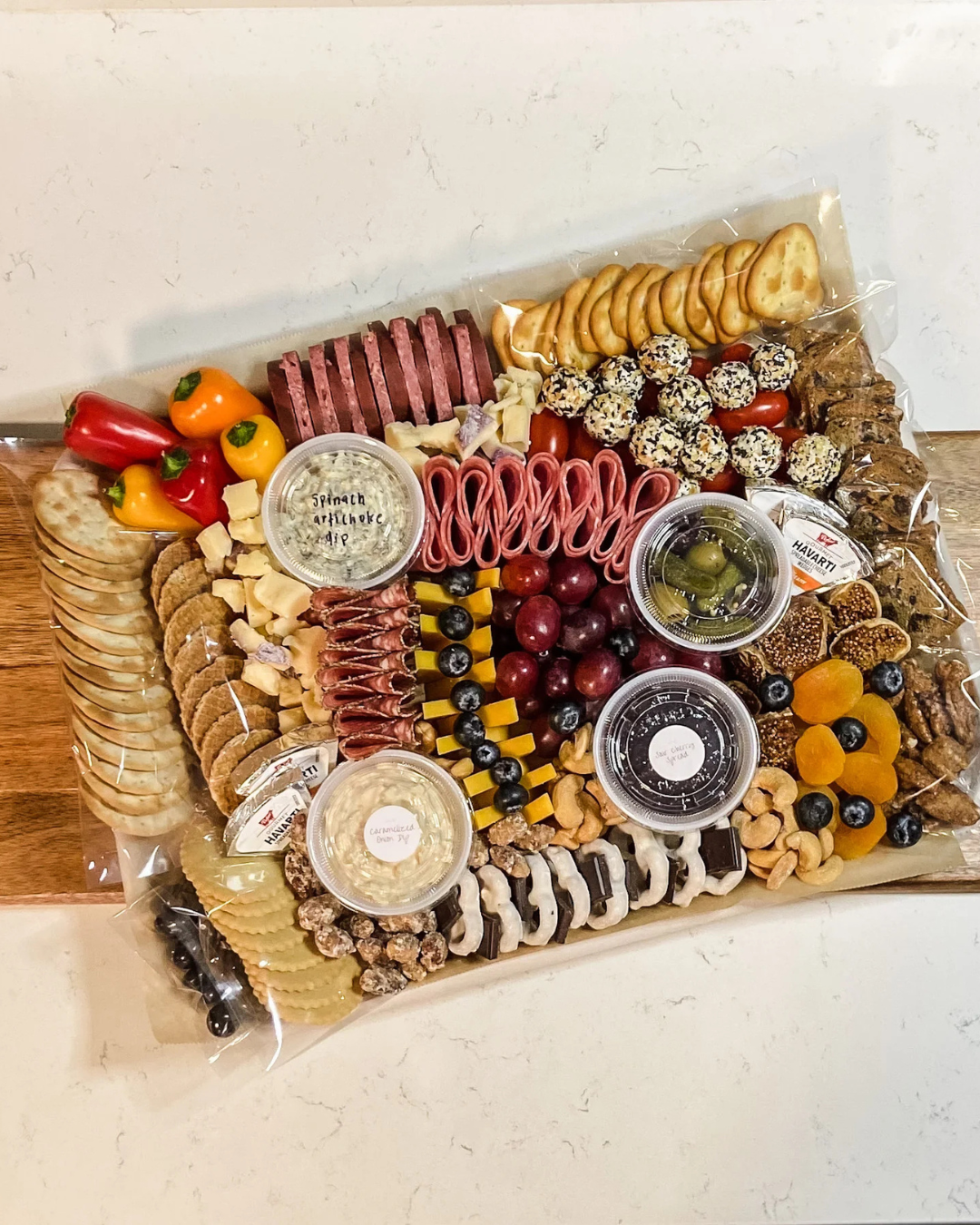 Large cheese and charcuterie board with assorted crackers, cheese slices, grapes, cherry tomatoes, pickles, nuts, dried fruits, hummus, and various meats, all arranged on a wooden surface.