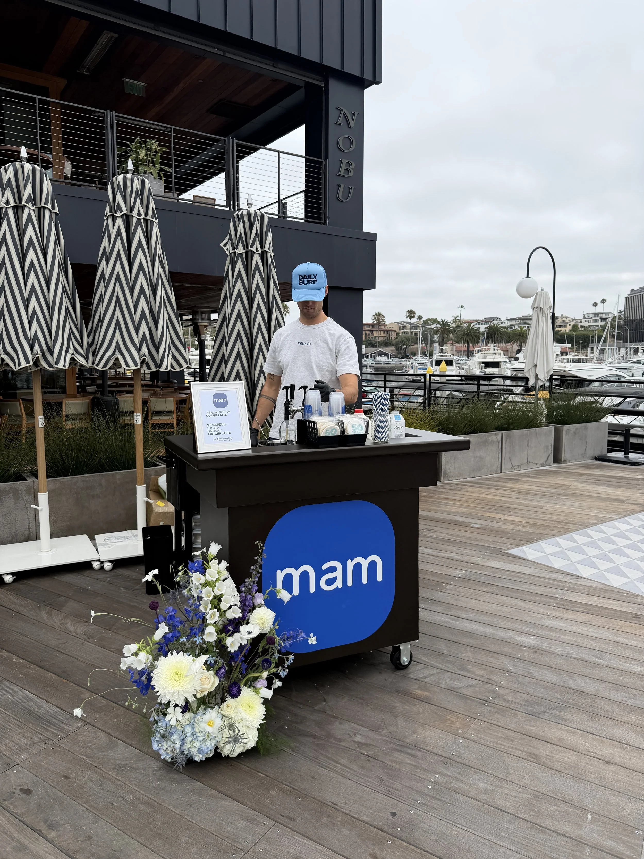 Coffee cart that is the color black as a blue square logo on the front with the word MAM in white lettering. A barista is pictured behind the coffee cart with an assortment of organic syrups and custom branded cups in front of him.