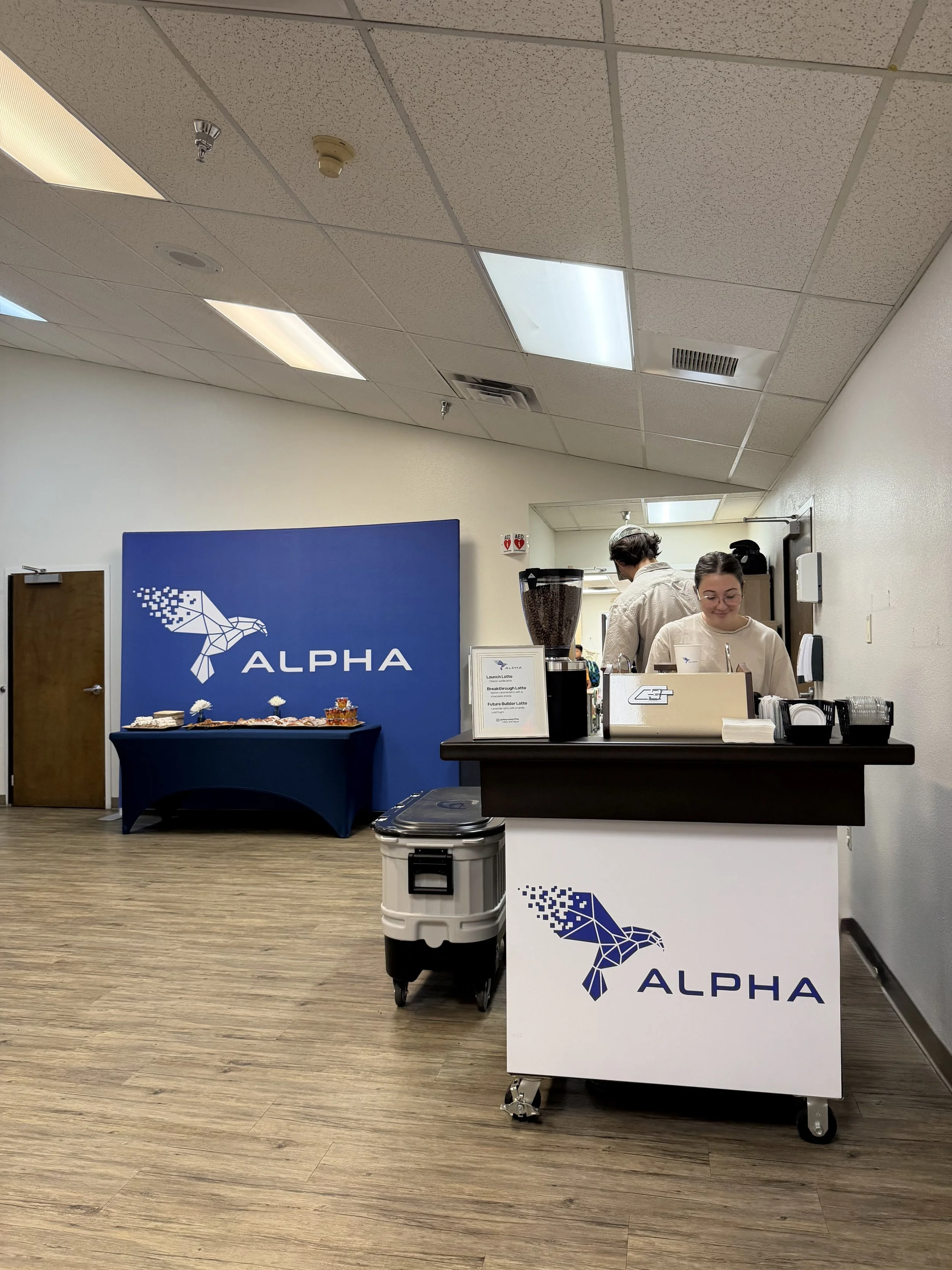 A coffee cart with a white front on it and a blue colored bird with the word "ALPHA" in blue lettering is pictured with two baristas behind the coffee cart and an espresso machine and other various coffee cart supplies.
