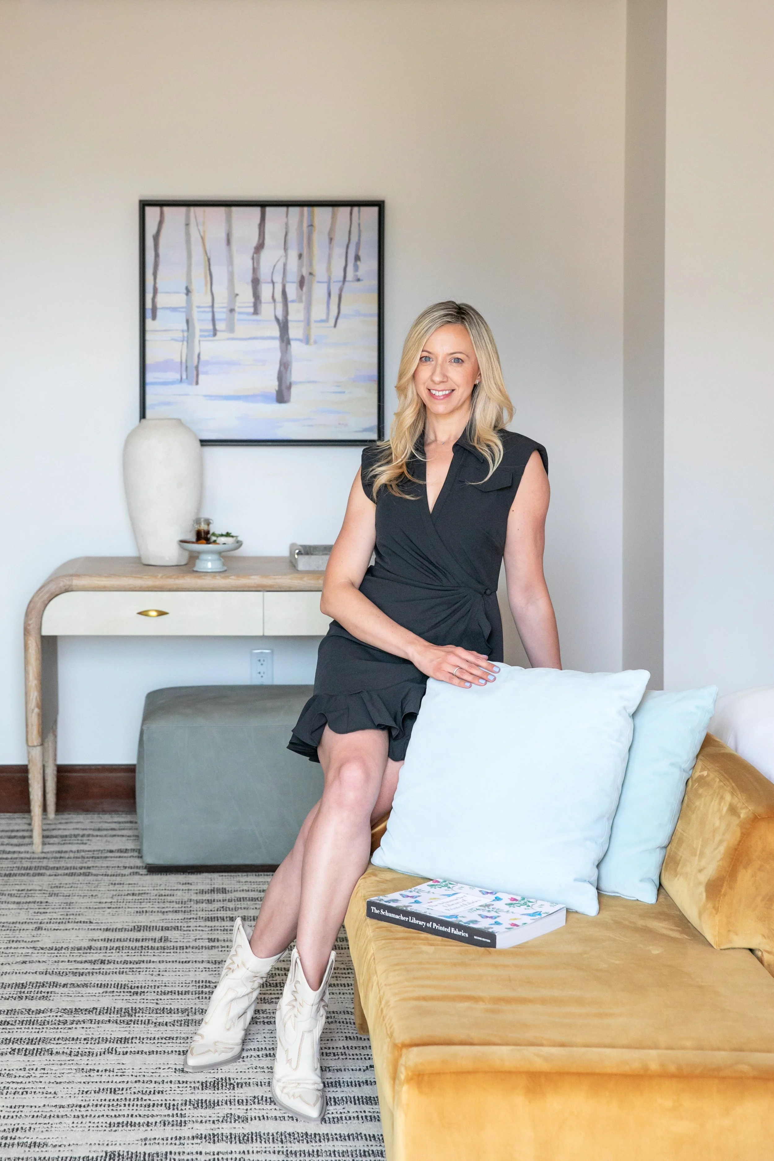 A woman with blonde hair in a black dress and white cowboy boots sitting on a yellow velvet couch with white and light green pillows, in a room with a white wall, a framed painting of trees, a white vase, a book, and a gray ottoman.