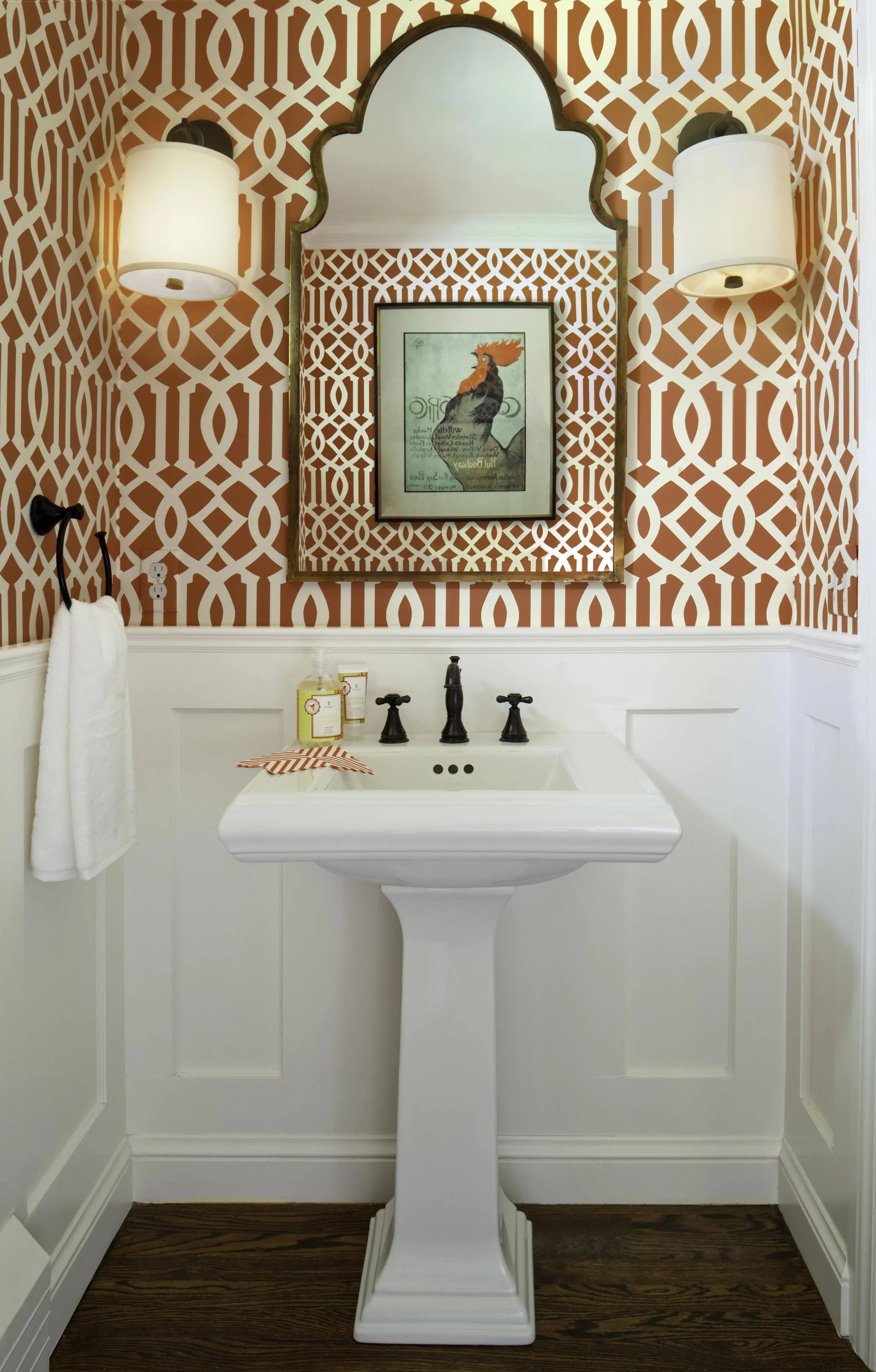 A bathroom with a white pedestal sink, a framed bird illustration above it, patterned red and white wallpaper, and wall-mounted lamps on either side of the mirror.