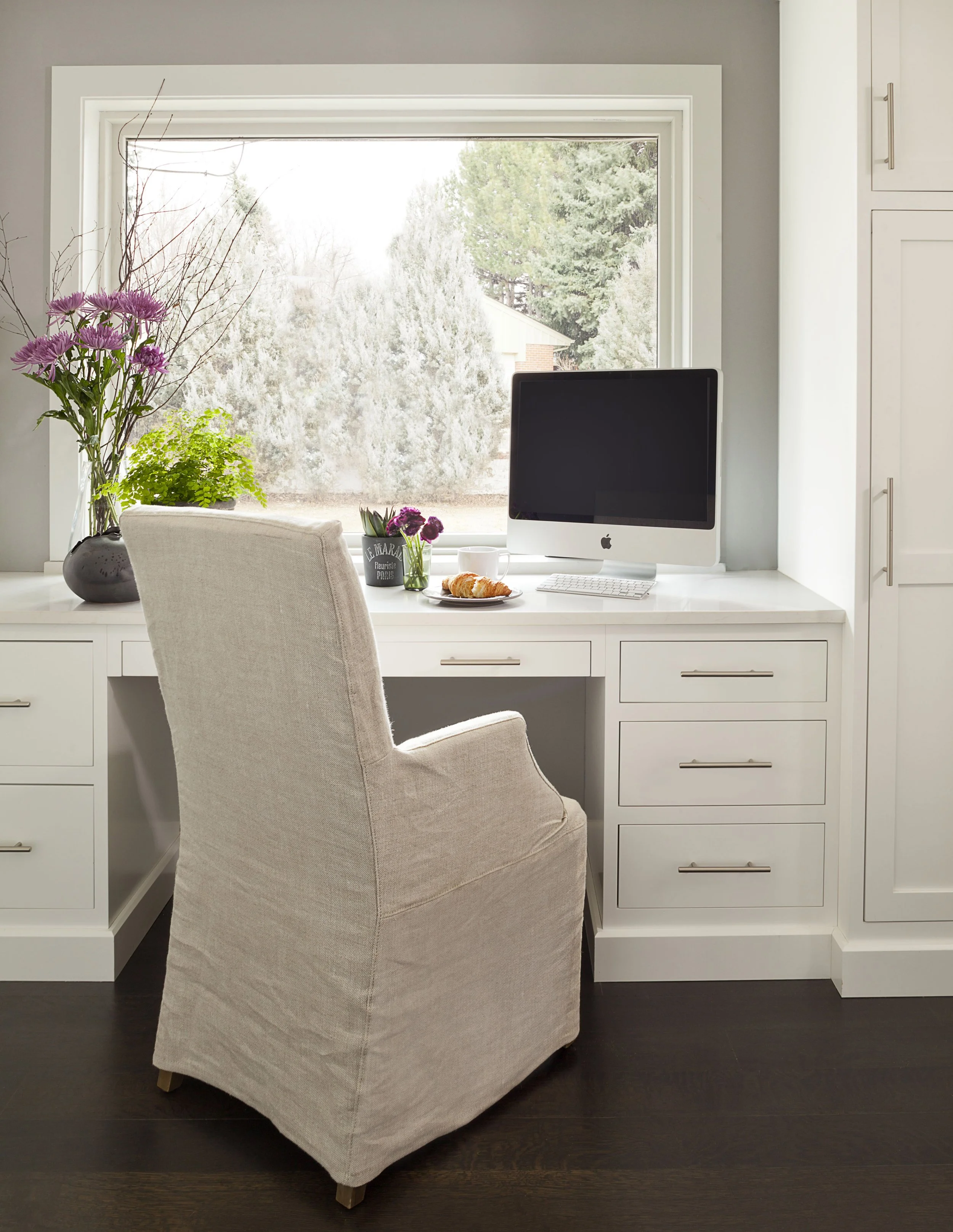 Home office workspace with a beige upholstered chair, white desk, Apple computer, and decorative flowers, set against a large window overlooking a garden.