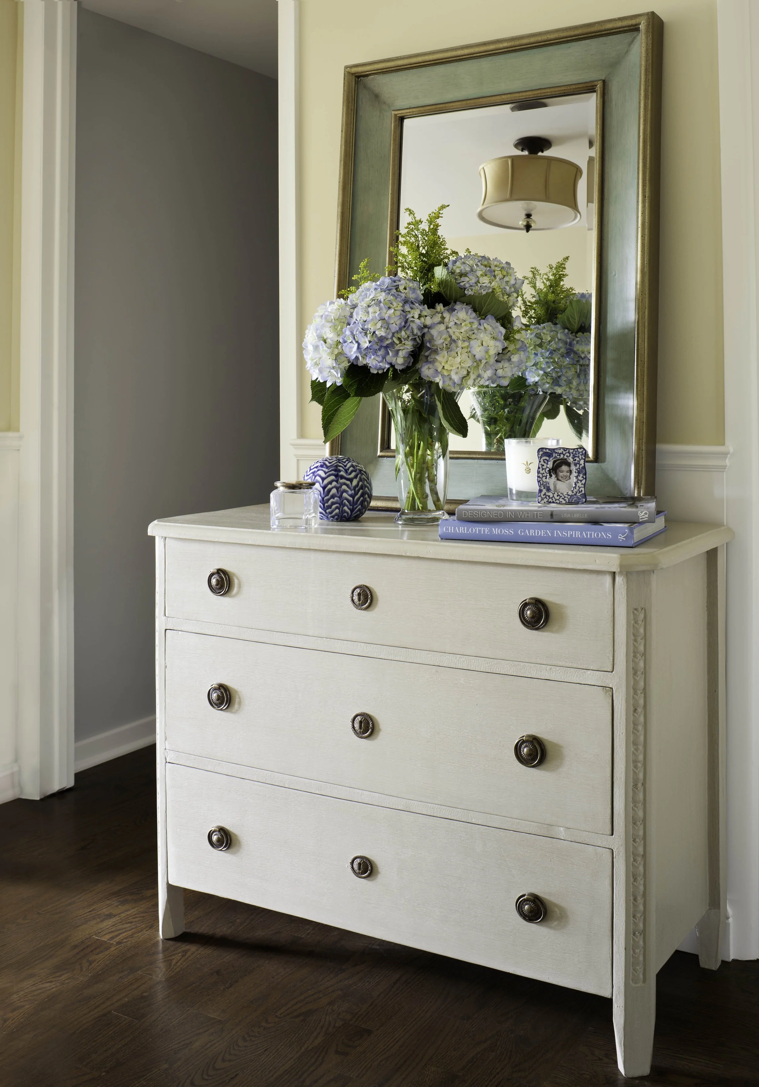 A cream-colored dresser with seven drawers and dark round knobs, topped with a flower arrangement of blue hydrangeas in a glass vase, a small decorative blue and white object, a candle, a stack of books, and a framed photo. A large mirror hangs above