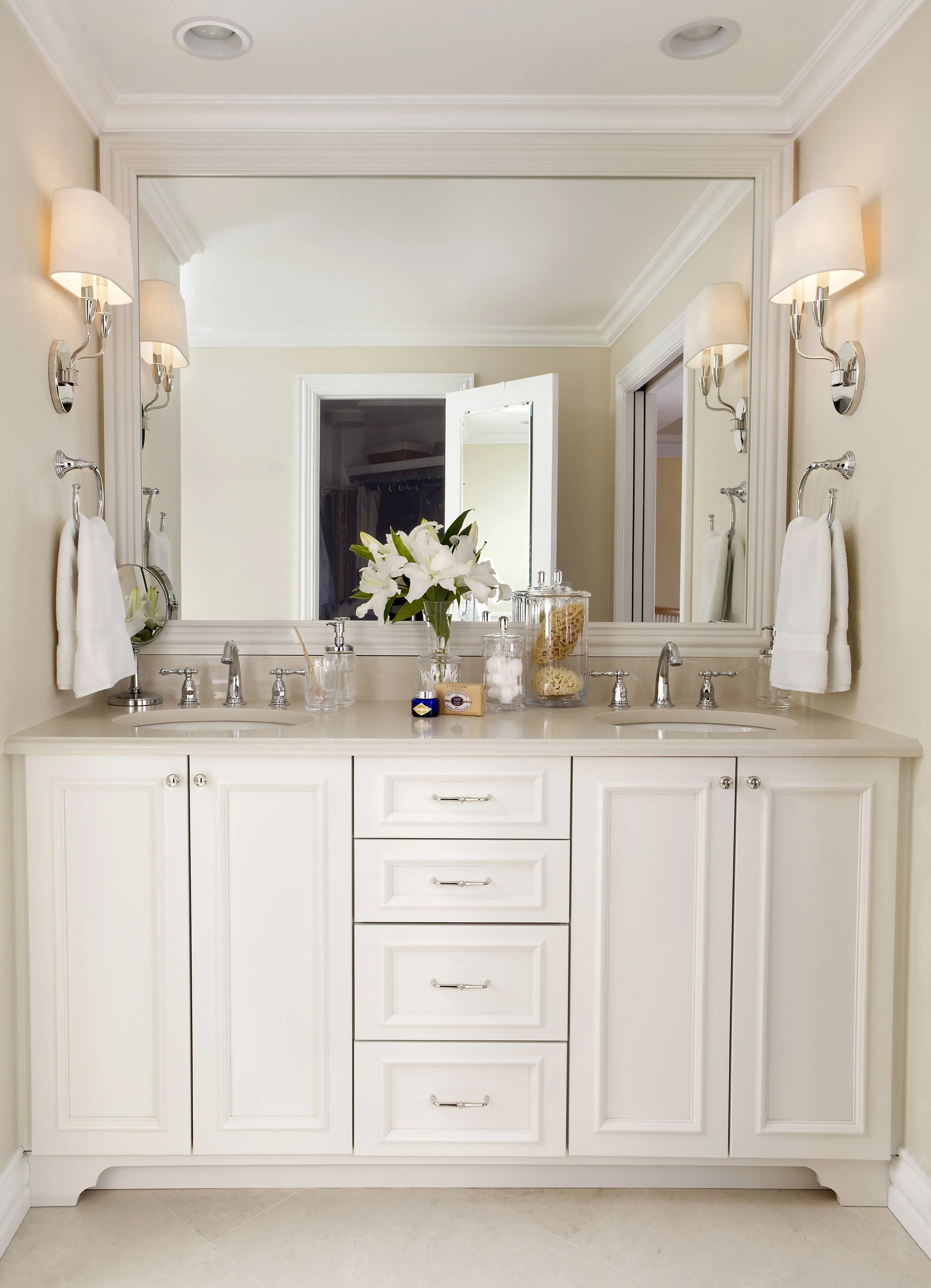 Bathroom vanity with two sinks, large mirror, white wooden cabinets, decorative items, flower vase with white lilies, wrapped towels, wall sconces, in a bright, neutral-colored bathroom.