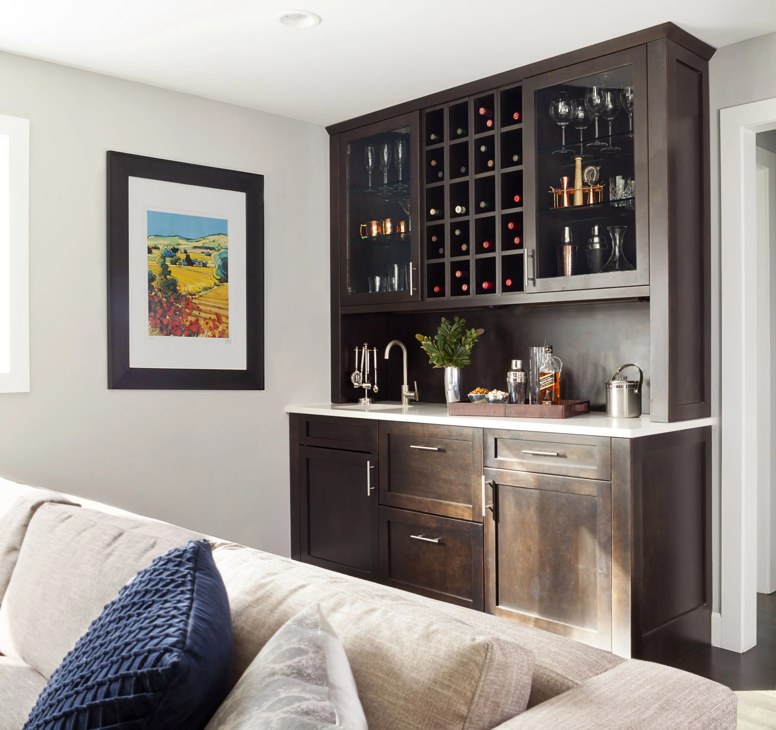 A home bar area with dark wood cabinets, glassware, and wine bottles, adjacent to a beige sofa with blue and white pillows, and a colorful landscape painting on a light gray wall.