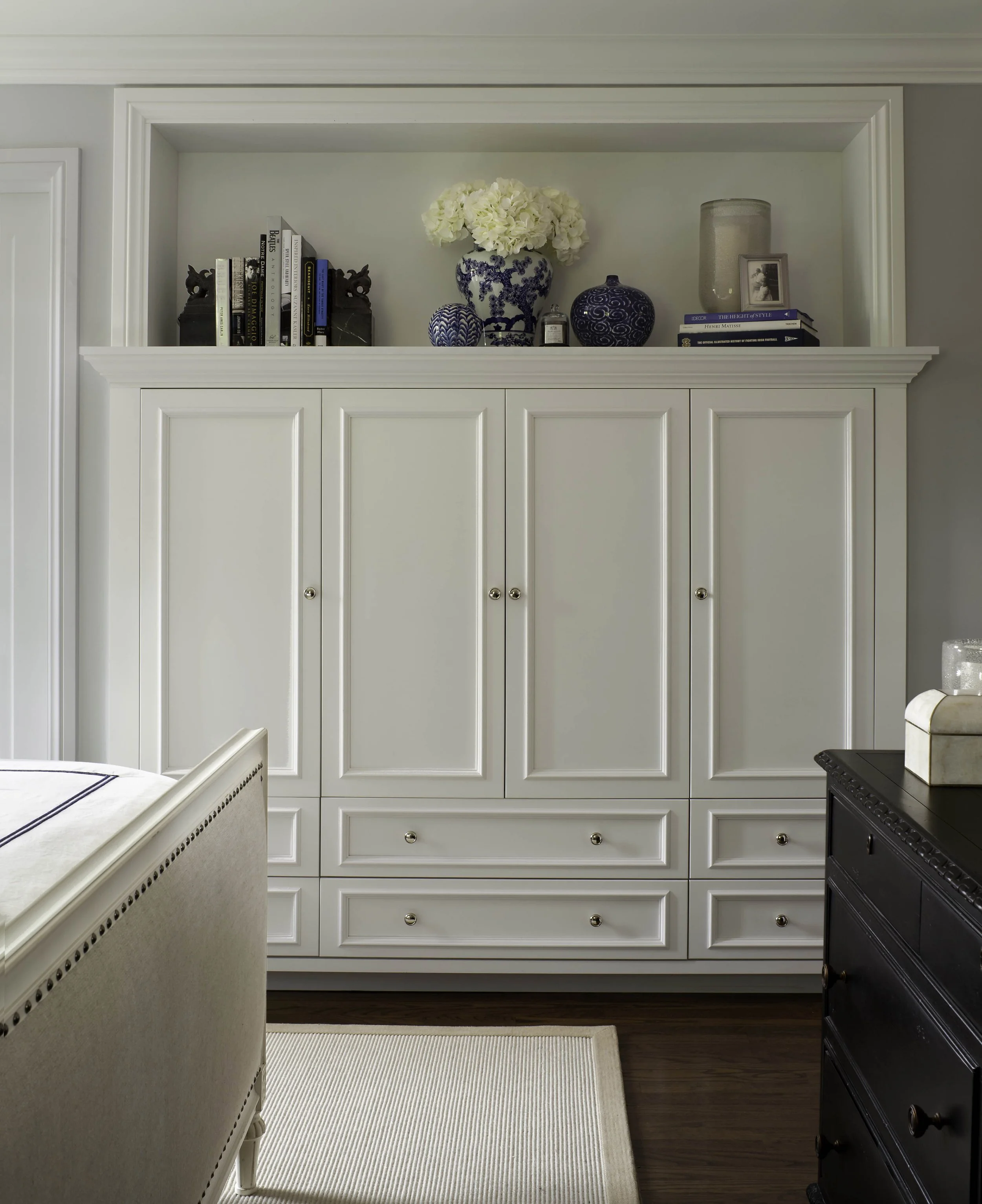 White wooden cabinet with framed details, three upper doors, and four lower drawers, situated in a room with a beige area rug and dark wood floor. Decorative blue and white vases, a flower arrangement, books, and framed photos are on top of the cabin