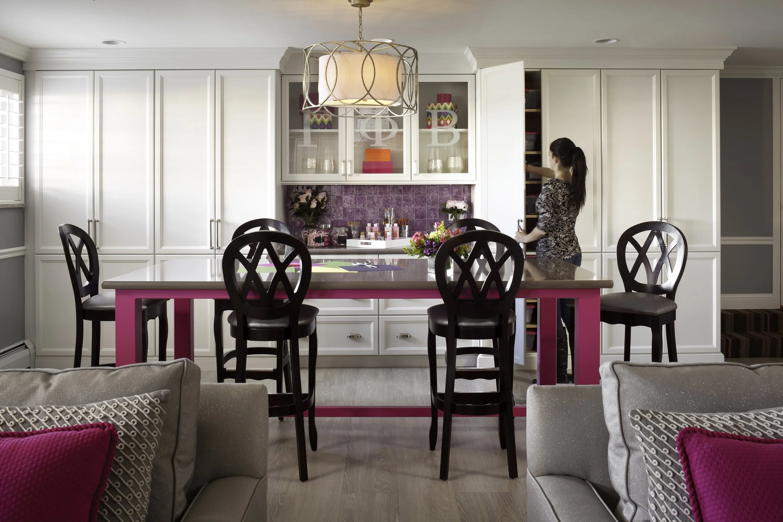 A woman organizing items in a storage cabinet in a bright kitchen with white cabinets, a pink and purple backsplash, a floral centerpiece on the counter, and a pink and gray kitchen island.