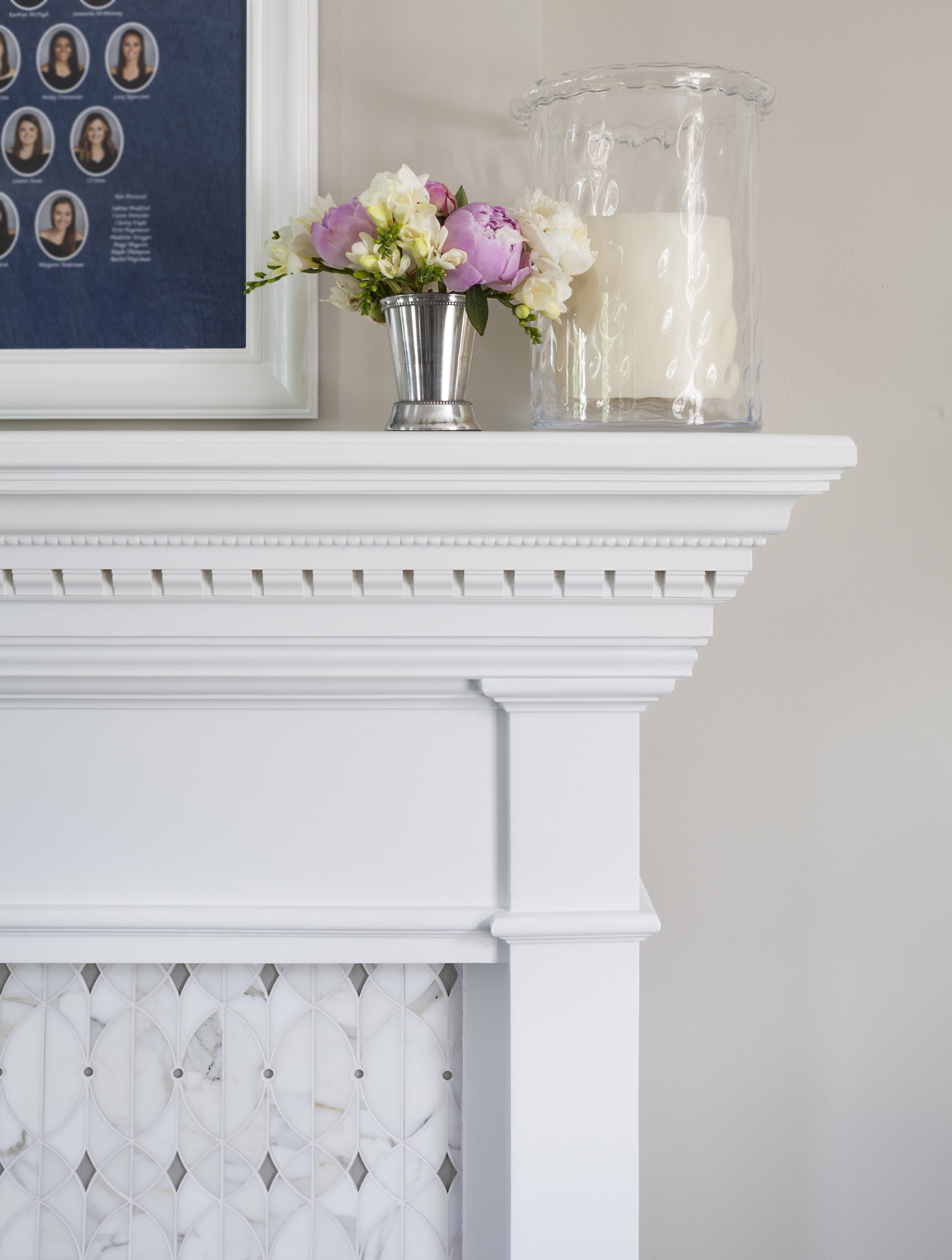 Decorative white fireplace mantel with a bouquet of pink and white flowers in a silver vase, a large glass hurricane candle holder with a candle, and a framed photo collage on the wall.