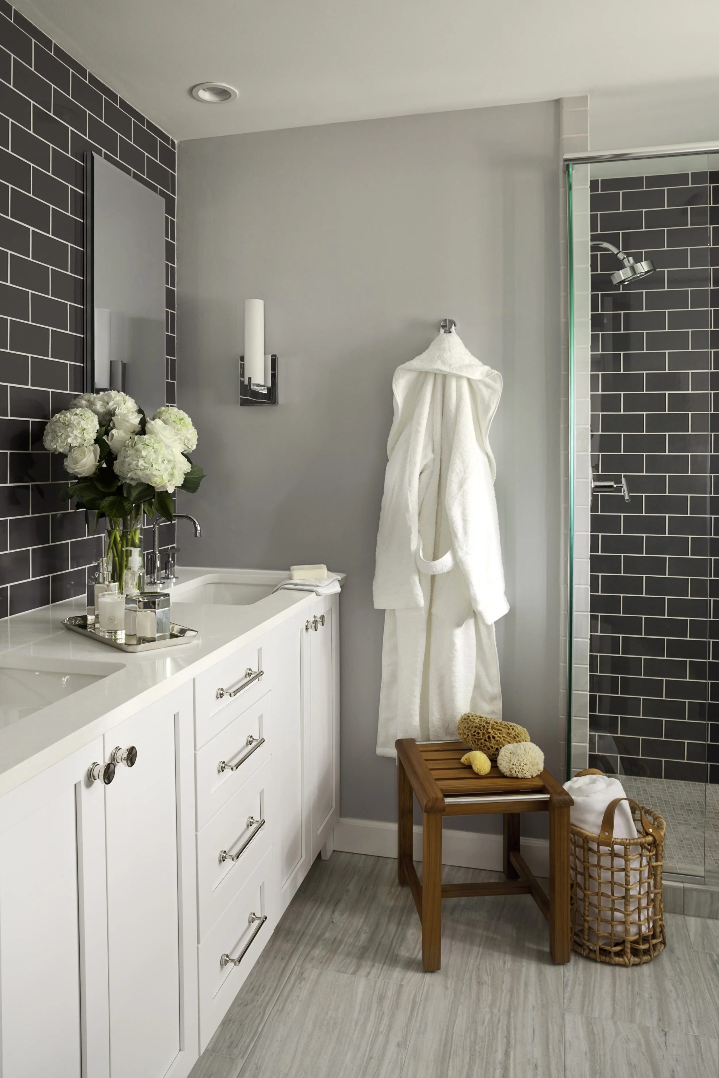 A modern bathroom with a white vanity, a vase of white flowers, a soap tray, a hanging white bathrobe, a small wooden bench with bath sponges, and a shower with dark gray tiled walls.