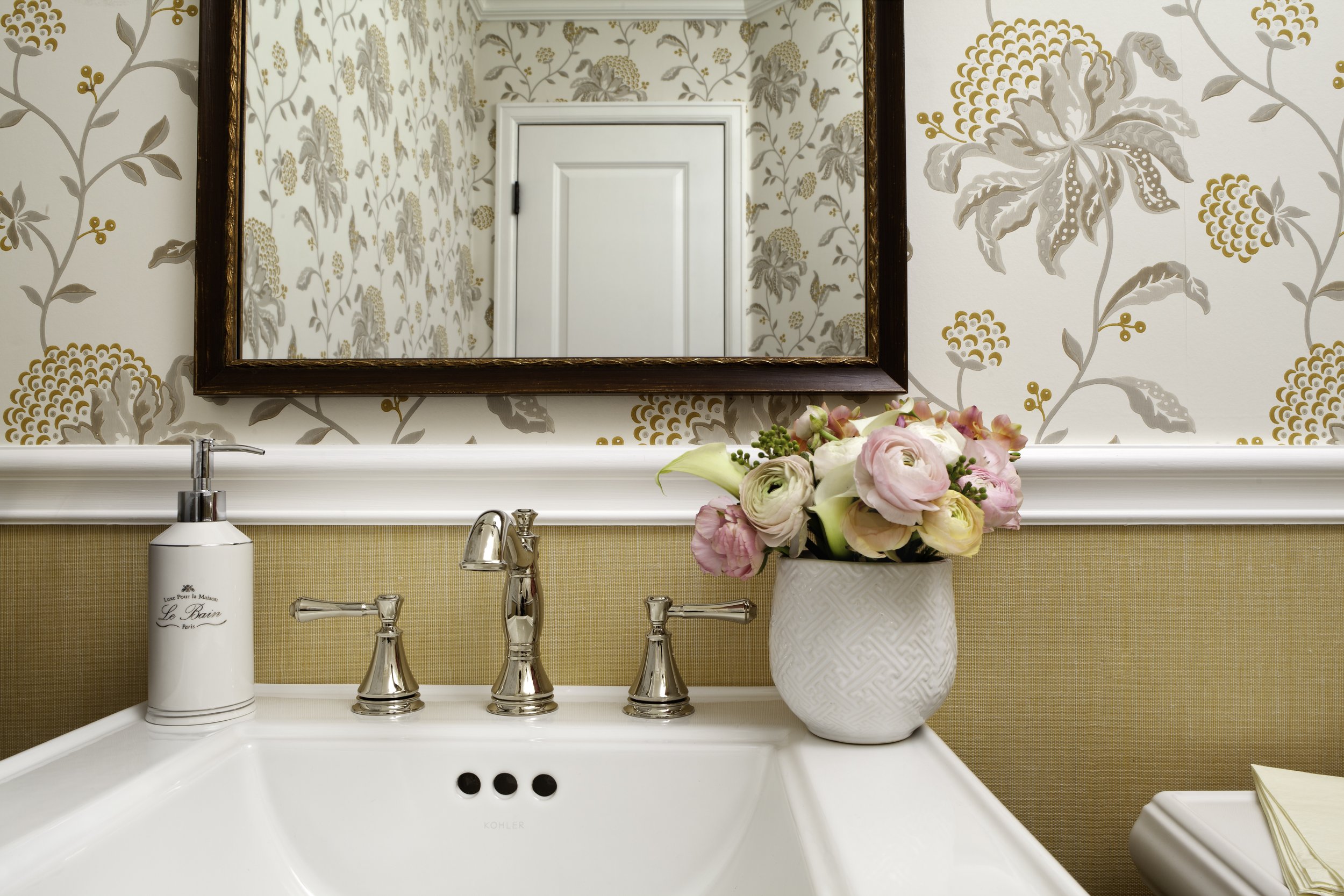 Bathroom sink with a white vase of pink and white flowers, a soap dispenser, and a mirror above the sink reflecting a white door with floral wallpaper.