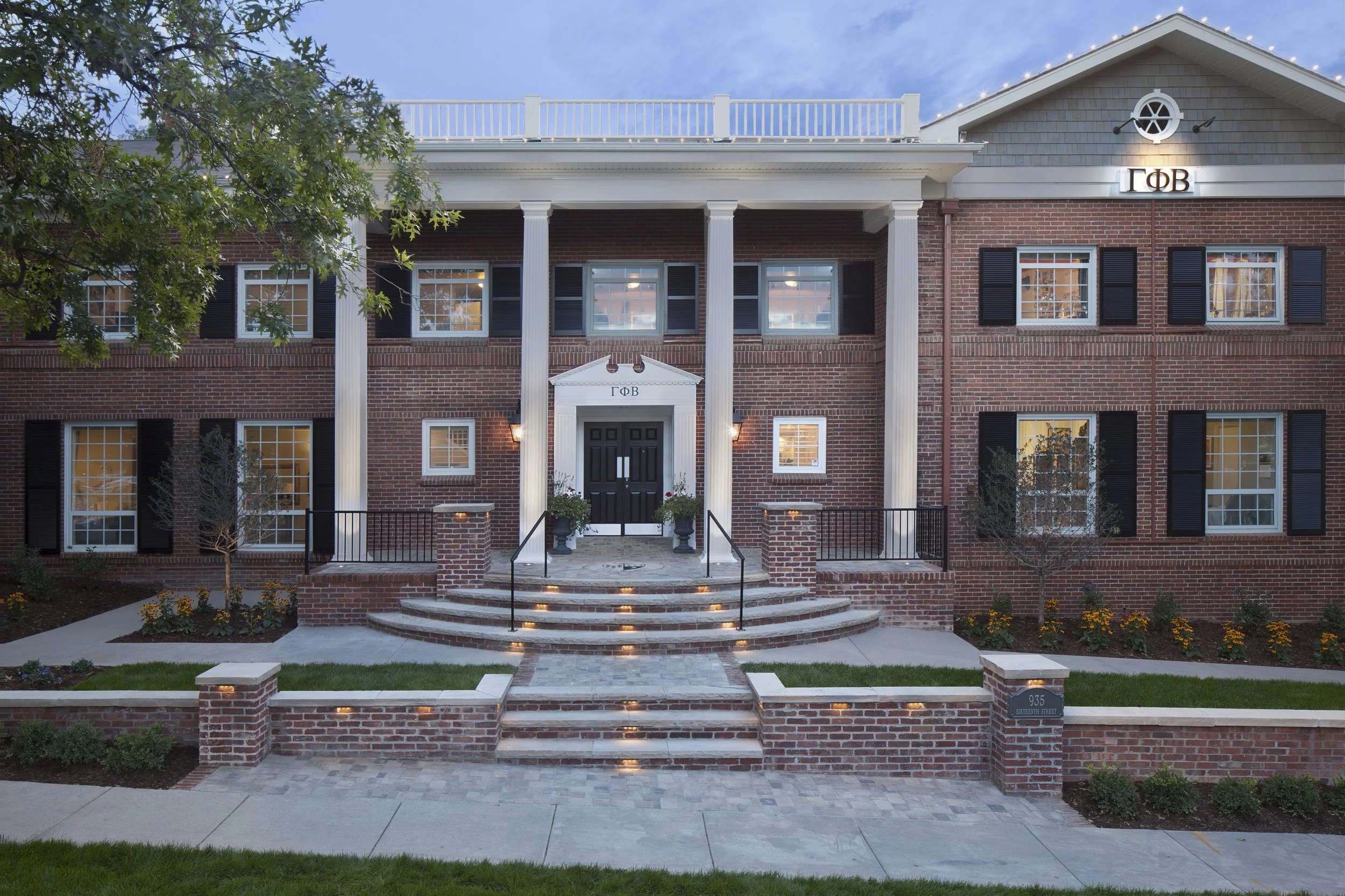 Front view of a large brick house with white columns, black windows with shutters, a black door, and a small front garden with flowers and trees. The house is decorated with string lights.