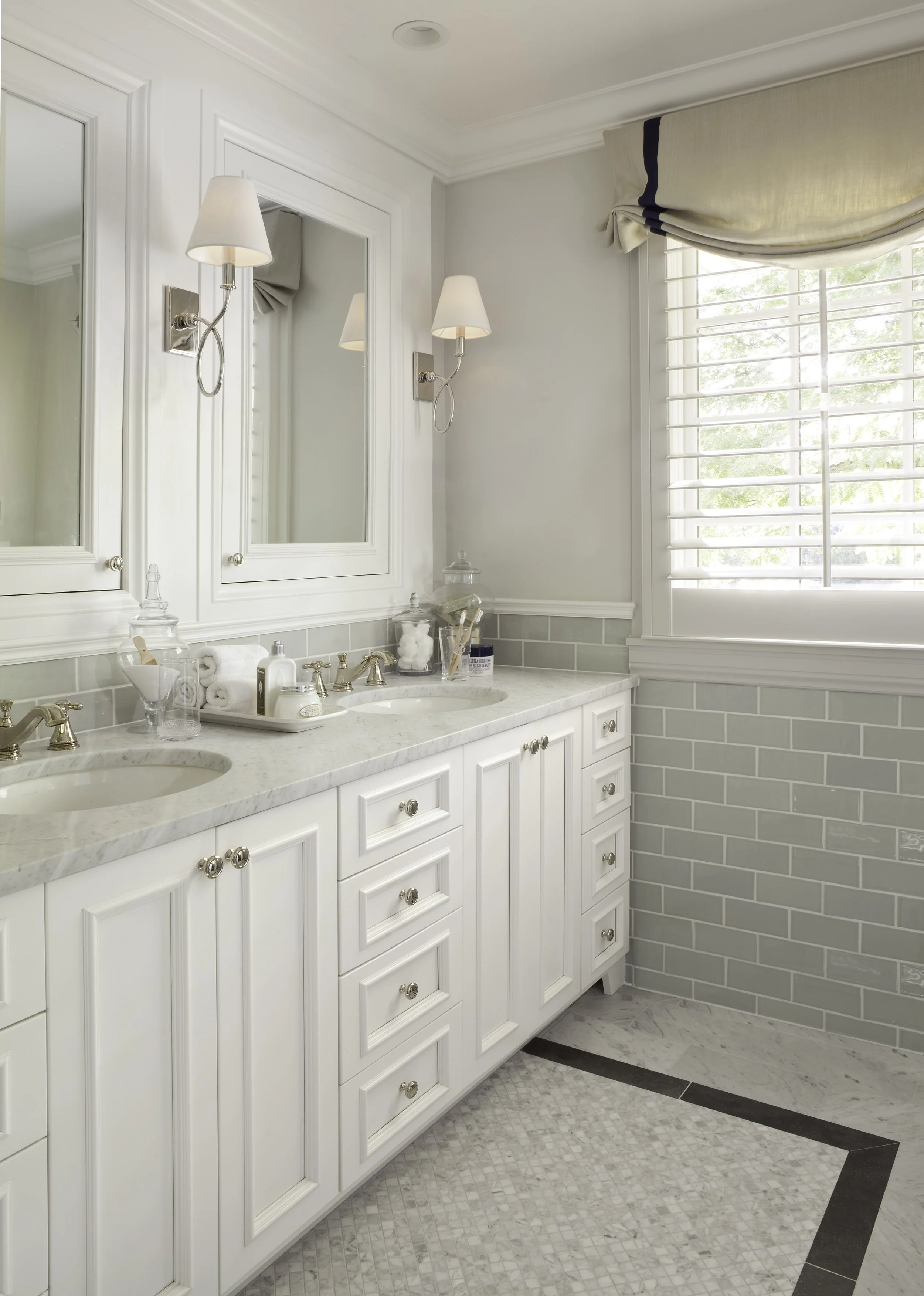 A bright, elegant bathroom with a white double vanity with marble countertop, mirrored medicine cabinets, wall-mounted light fixtures, a window with white plantation shutters, and light-colored tiled walls and floor.