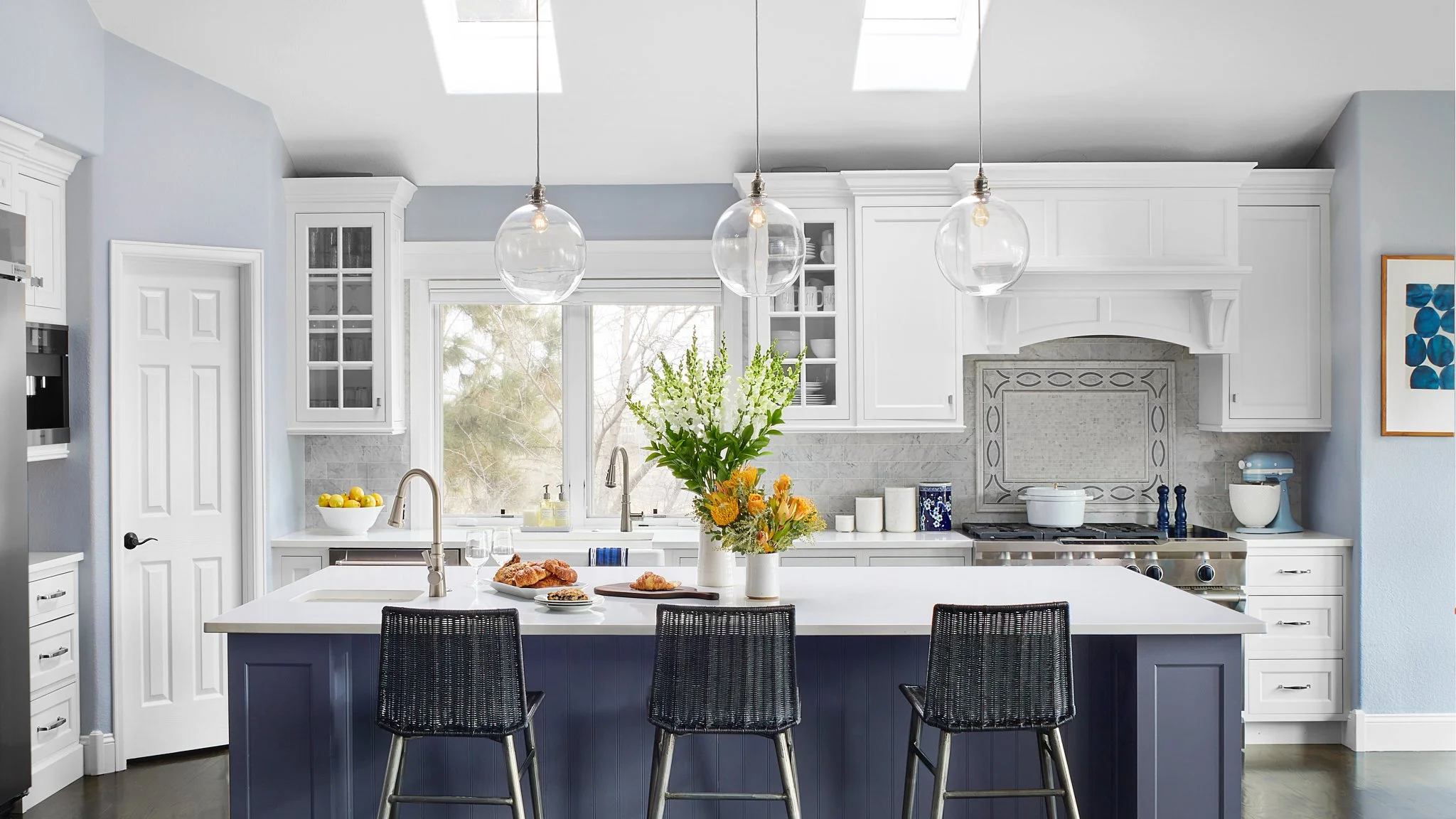 Modern kitchen with white cabinets, a navy blue island, three black bar stools, a large bouquet of flowers on the island, and skylights.