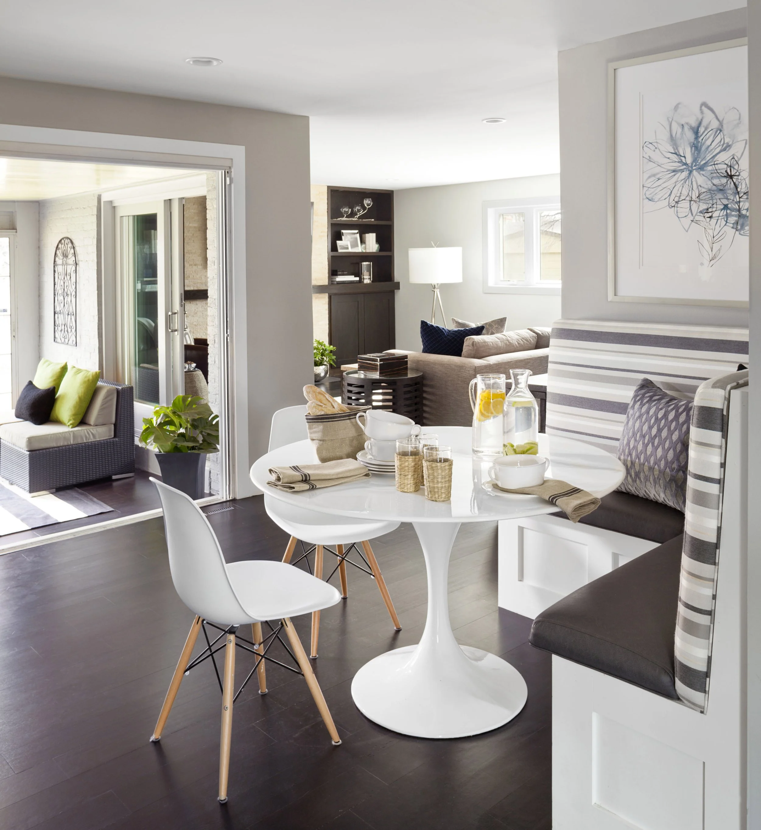Open-concept living and dining area with a round white table, white chairs, built-in bench seating with striped and patterned cushions, and a cozy sitting area with a beige couch, coffee table, and window in a modern home.