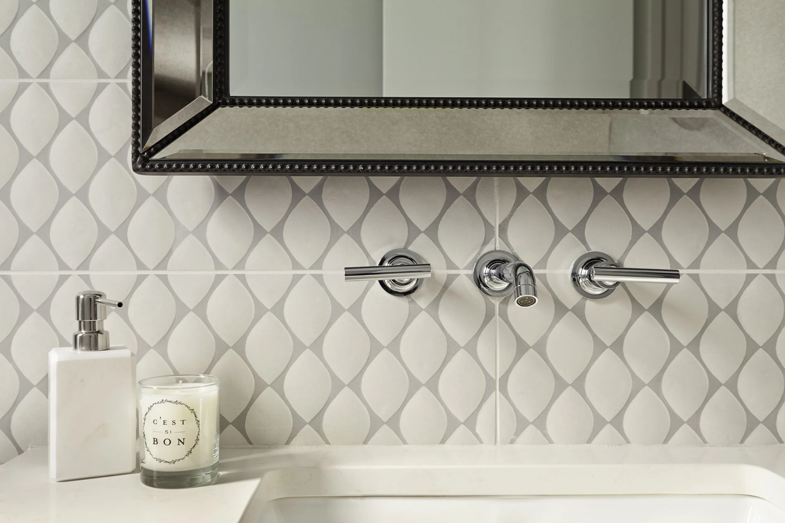 Bathroom mirror above a sink with a soap dispenser and a candle on the countertop, patterned tile wall.