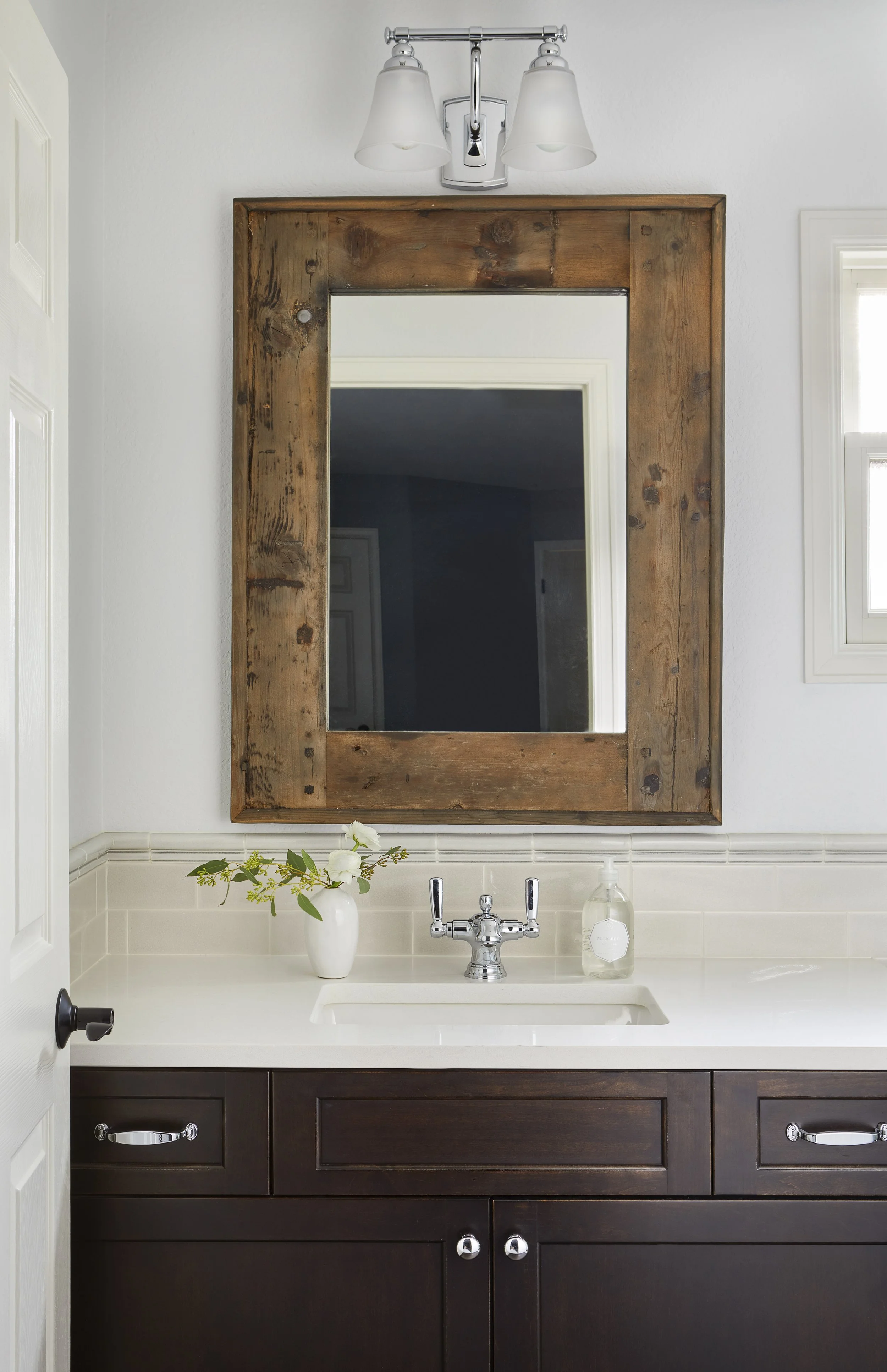 Bathroom sink with dark wood cabinet, white countertop, and a large wooden framed mirror. Small vase with green leaves and white flowers, soap dispenser, and wall-mounted light fixture above the mirror.
