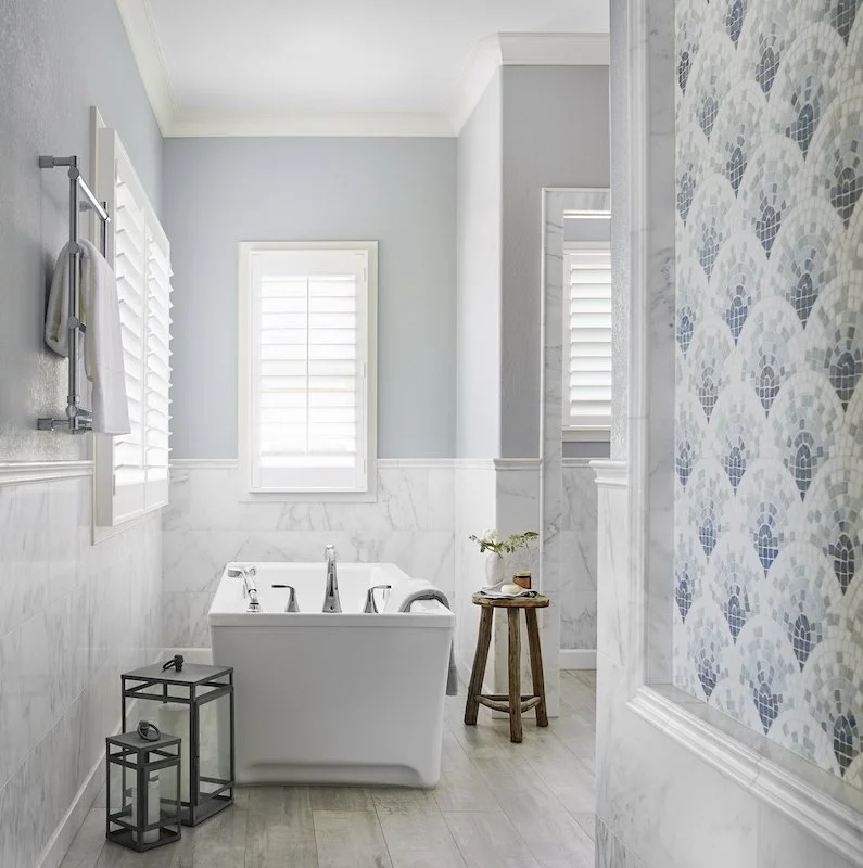 Bright bathroom with a white bathtub, two window shutters, a small wooden stool with a plant and soap, decorative candlesticks, and patterned wall tiles.