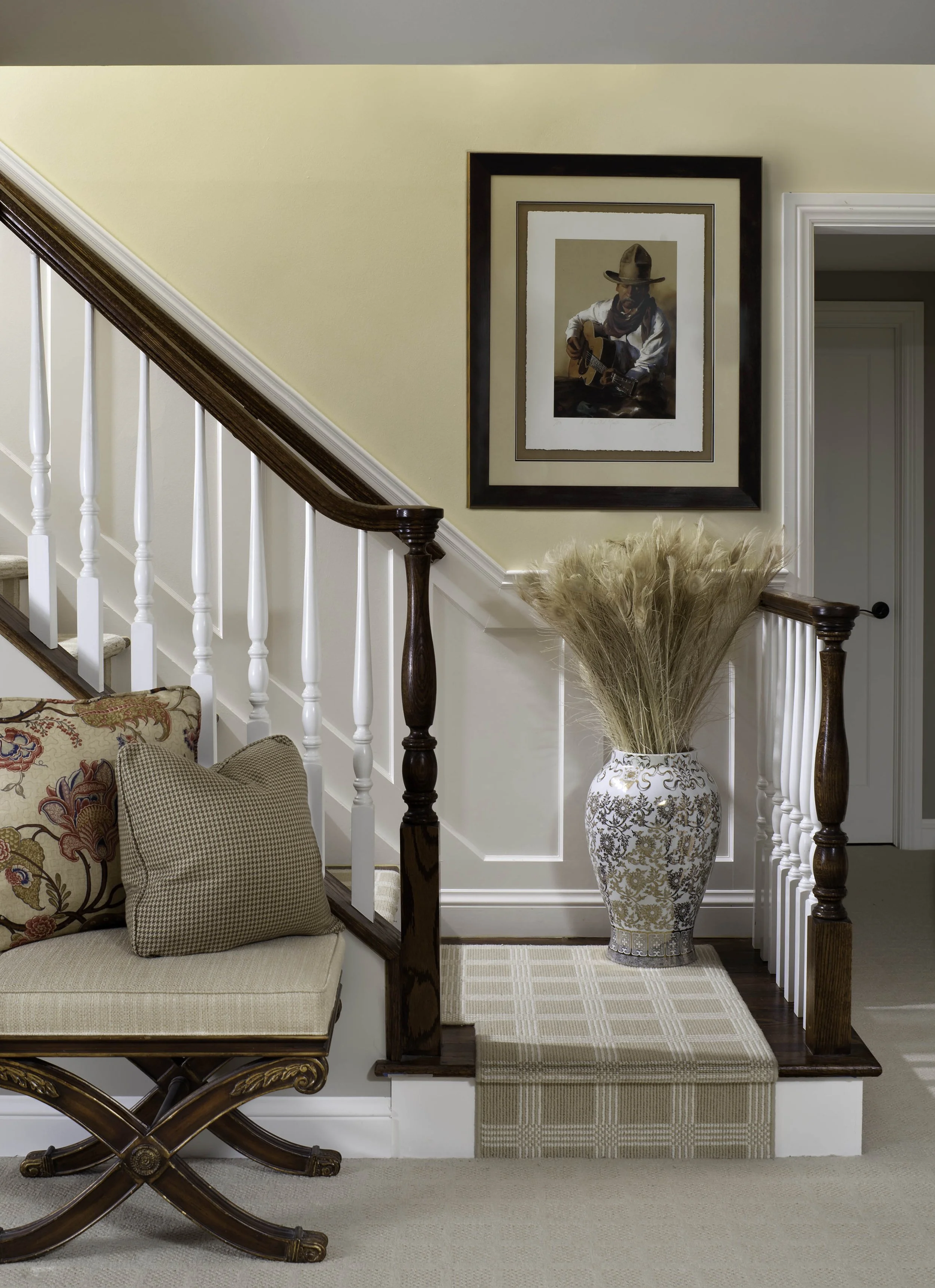 A staircase landing with a large decorative vase filled with pampas grass, a framed picture of a man playing guitar, a cushioned wooden bench, and beige carpeting.