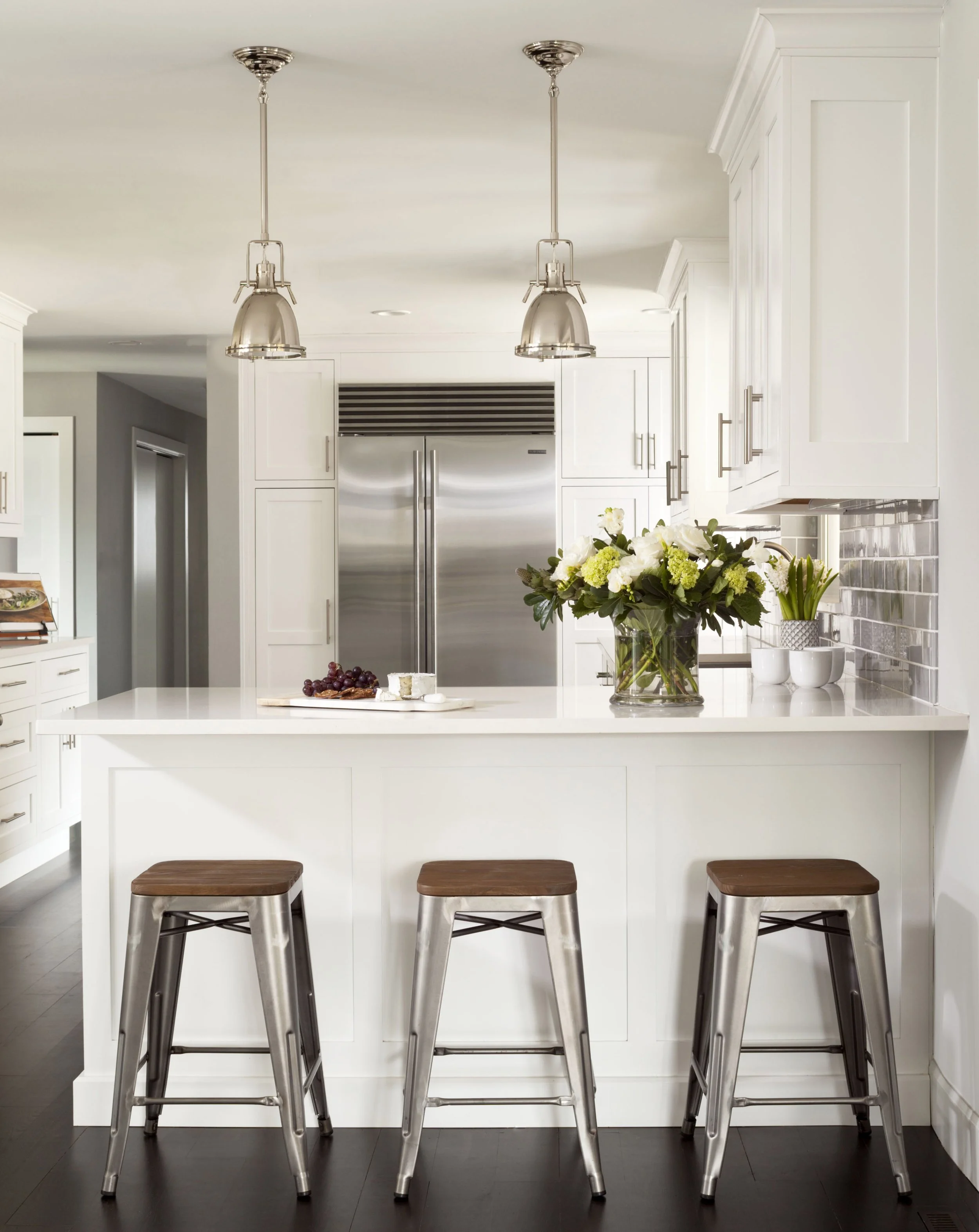Modern white kitchen with a large island, three metal barstools with wooden seats, stainless steel refrigerator, vase of flowers, and pendant lighting.
