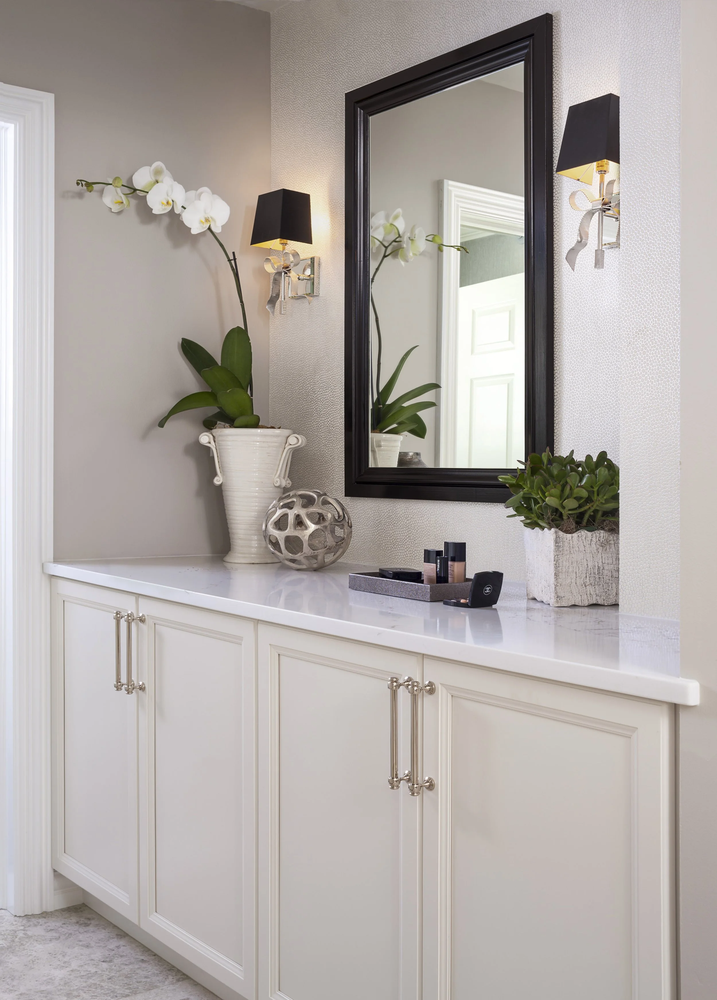 A white bathroom vanity with a marble countertop, decorated with a potted orchid, a decorative ceramic sphere, and a lotion tray with makeup bottles. A large rectangular mirror with a black frame hangs above, flanked by black wall sconces with ribbon