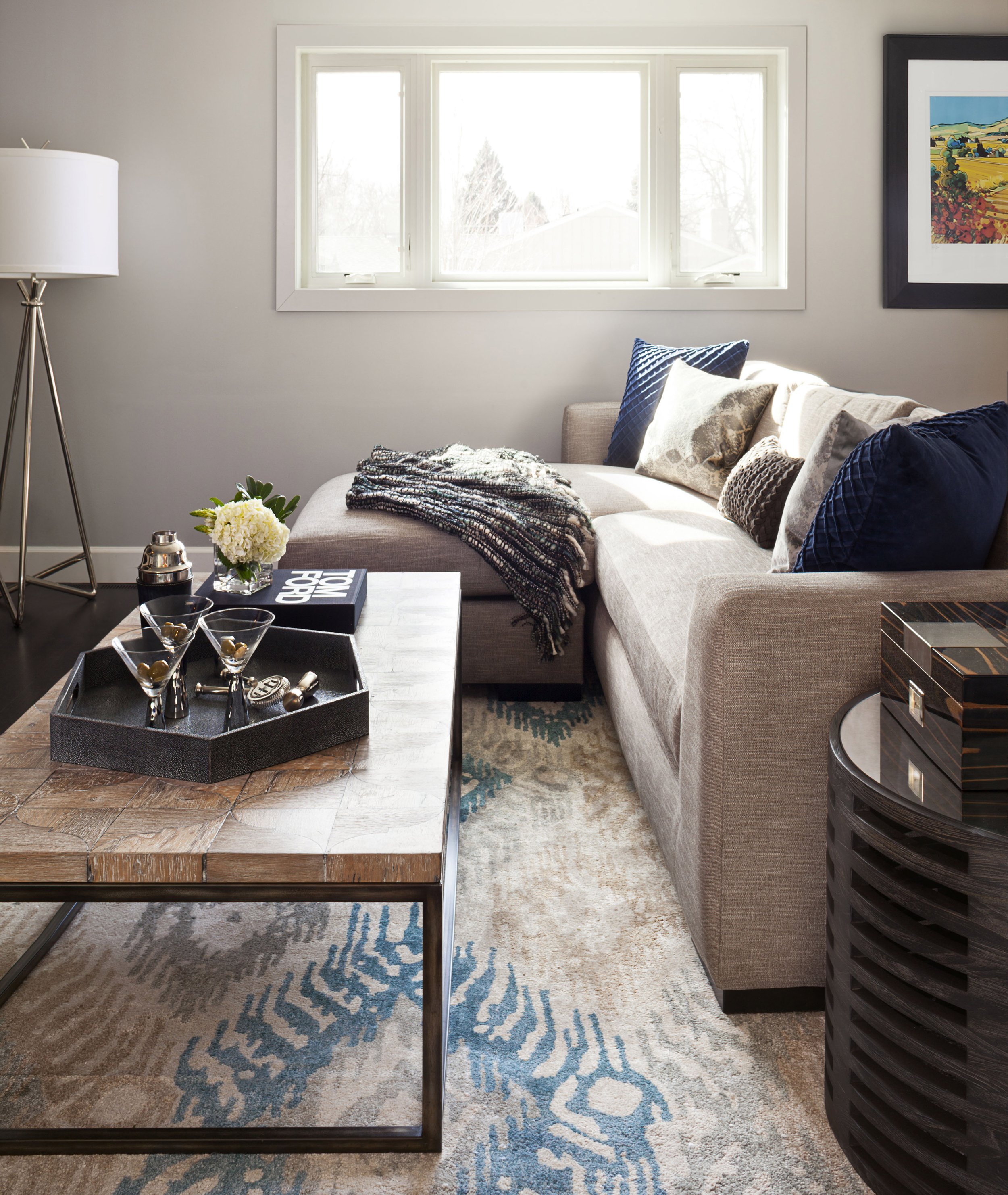 Living room with beige L-shaped sofa, decorative pillows, a black tray with glasses and decor, a coffee table, a rug, a window, and a framed painting on the wall.