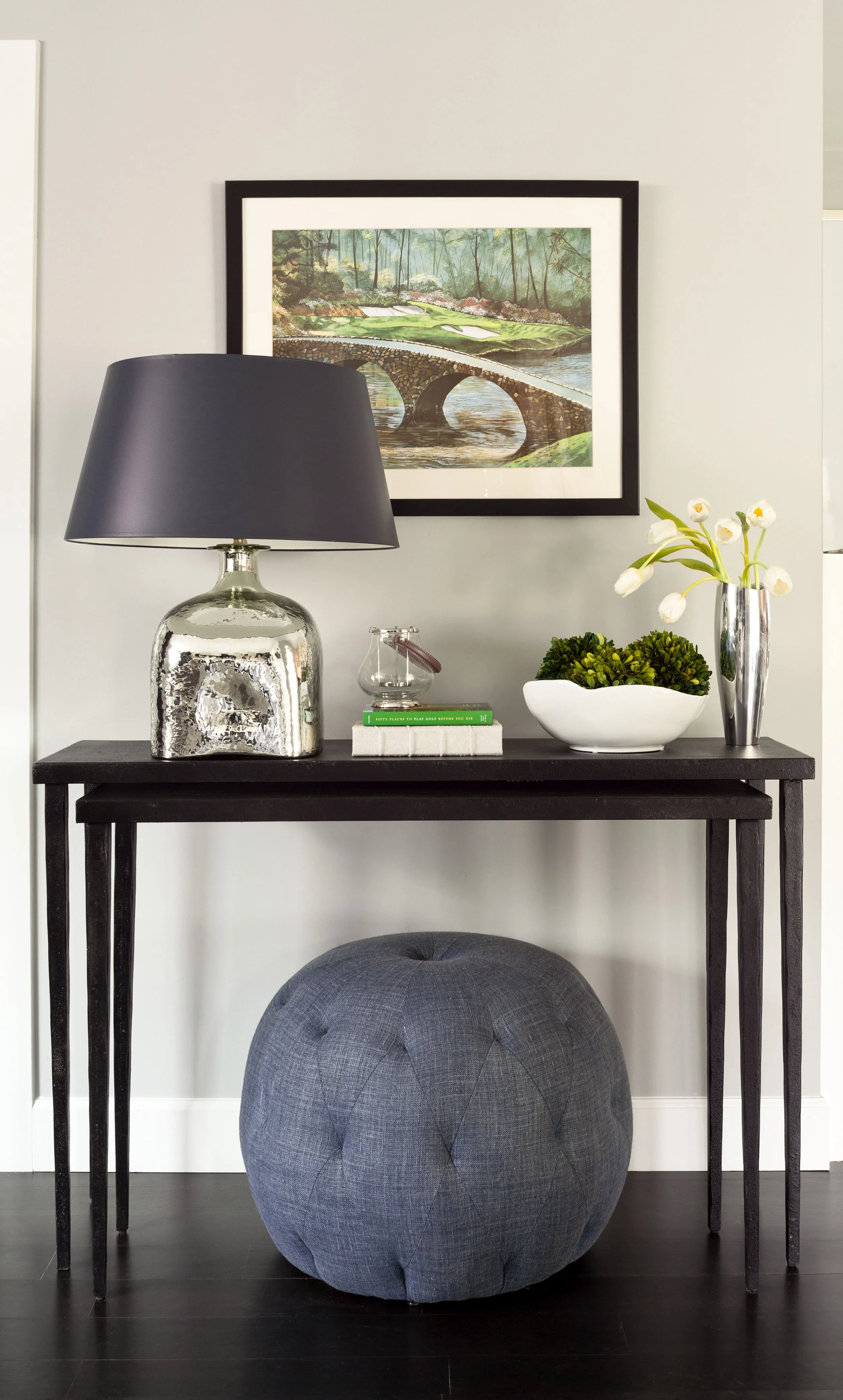 A black console table with a silver lamp, a glass container with a key, a stack of two books, a white bowl with greenery, and a tall silver vase with white flowers. A grey pouf sits in front of the table, and a framed landscape painting hangs on the 