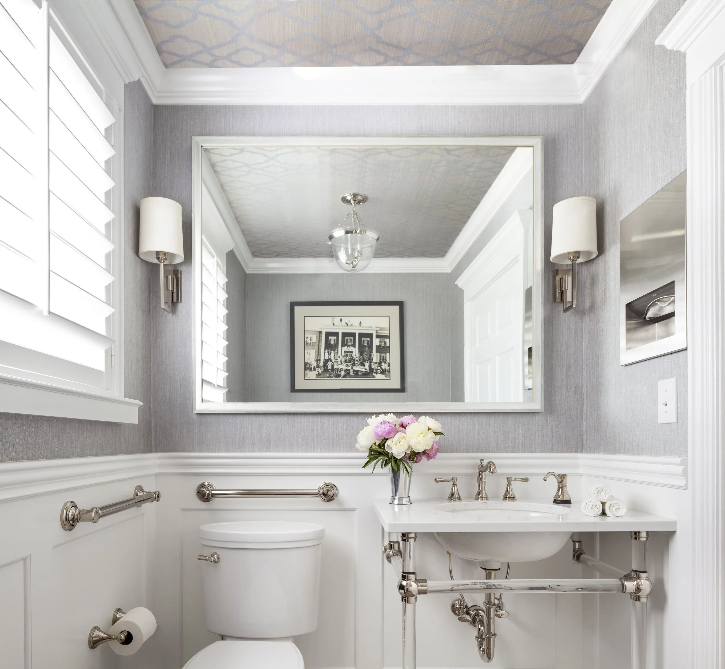 A small bathroom with a white toilet, a white sink with chrome fixtures, a large mirror, and a decorative ceiling. There are two wall-mounted light fixtures, a vase with pink and white flowers on the sink, and a rolled towel. Wainscoting and charcoal