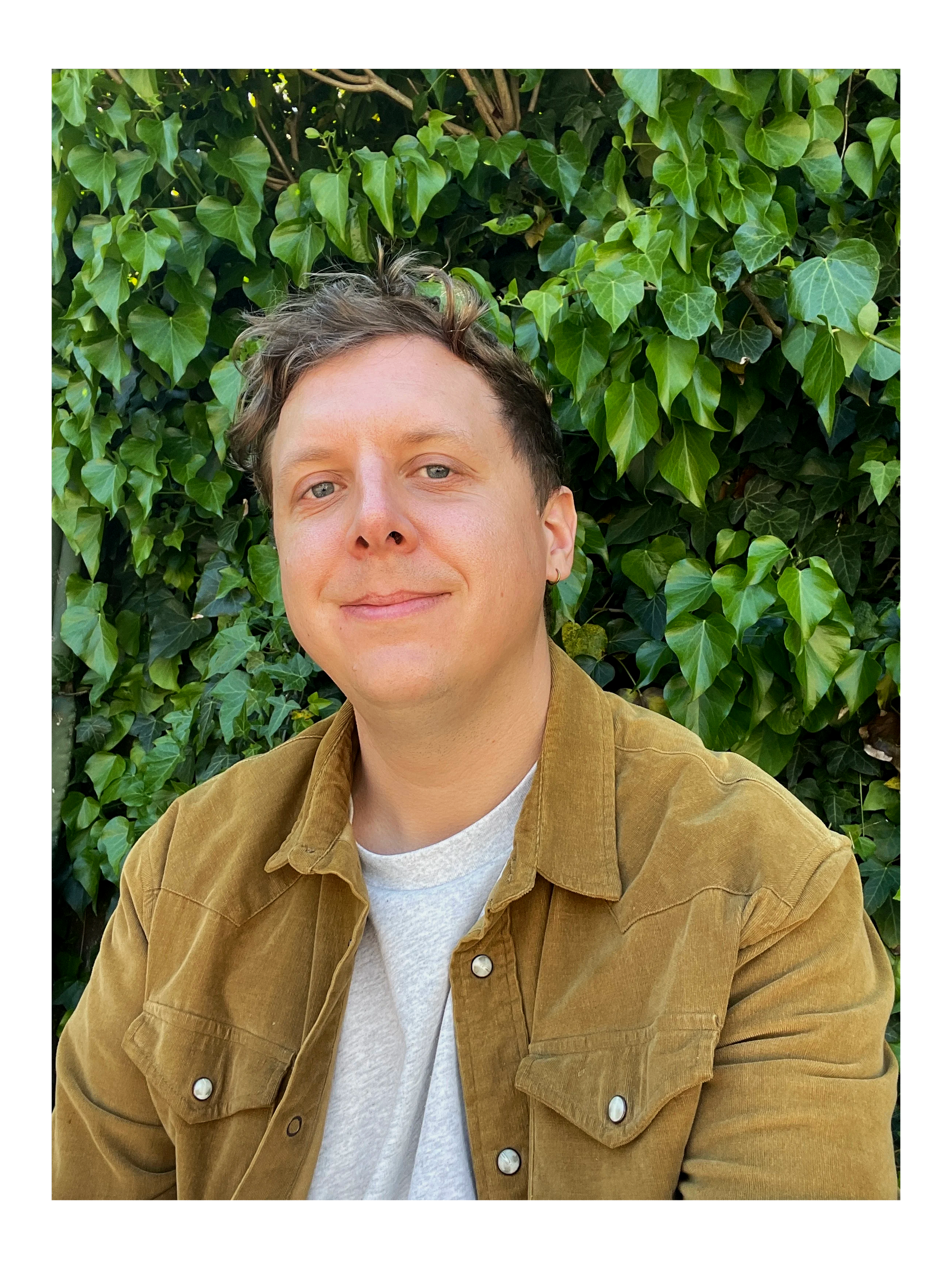 A person with short brown hair and light blue eyes, smiling slightly, wearing a tan jacket over a white shirt, standing outdoors in front of a wall of green ivy leaves.
