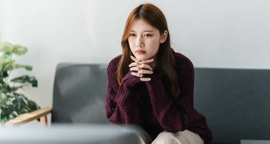 A young woman sitting on a couch with her hands clutched together.